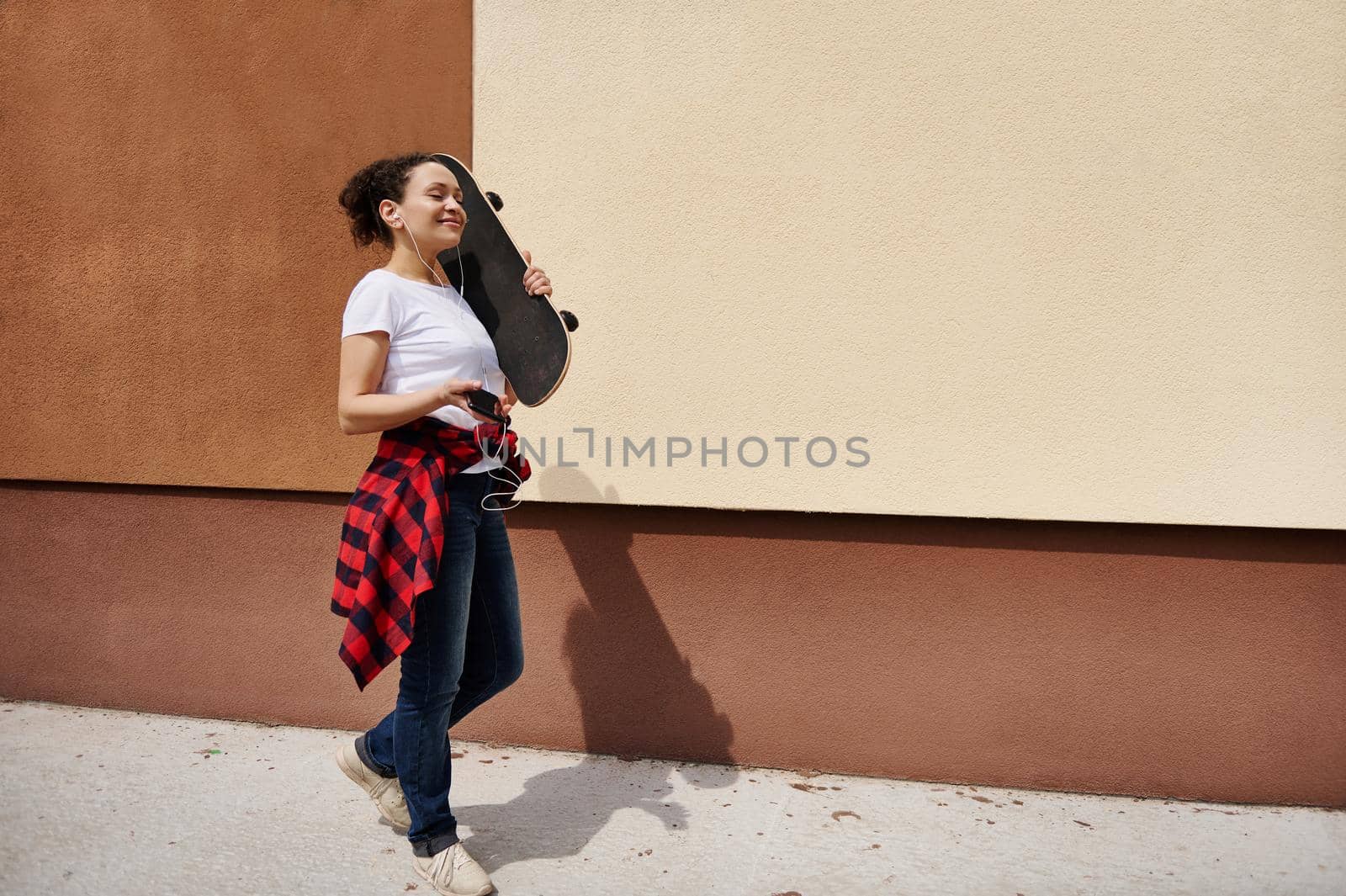 Happy hispanic woman holding wooden skateboard on shoulder walking down the street listening to music against colored wall background