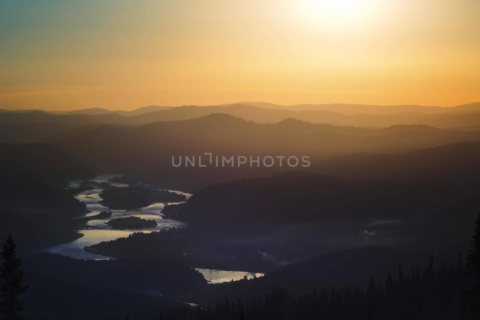 Golden sunset in the mountains: dark silhouettes of the hills, golden light in the haze, clouds in the blue sky, at the bottom of the valley reflection in the water of the river, Kuzbass Siberia