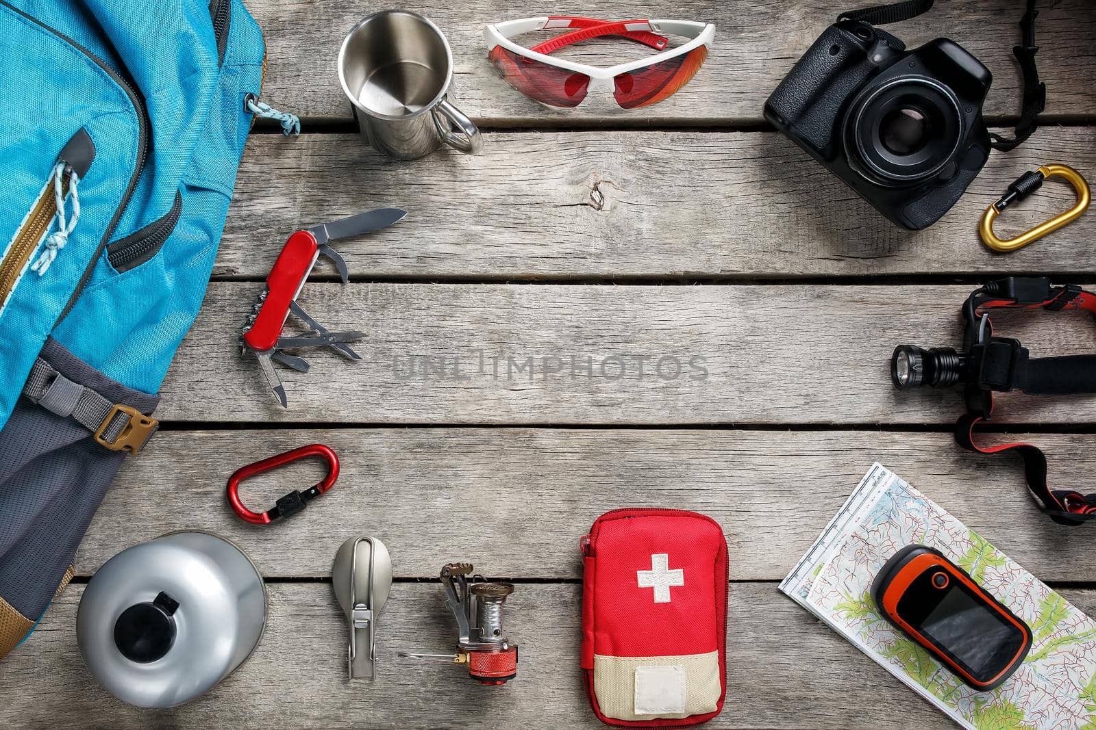 Top view of tourist equipment for travel and tourism on a rustic light wooden floor with an empty space in the middle. Items include a kettle, a card, a knife, a rope, a carbine, a flashlight, shoes, GPS, a gas bottle, a burner, a spoon, a mug, a camera, glasses, a backpack.