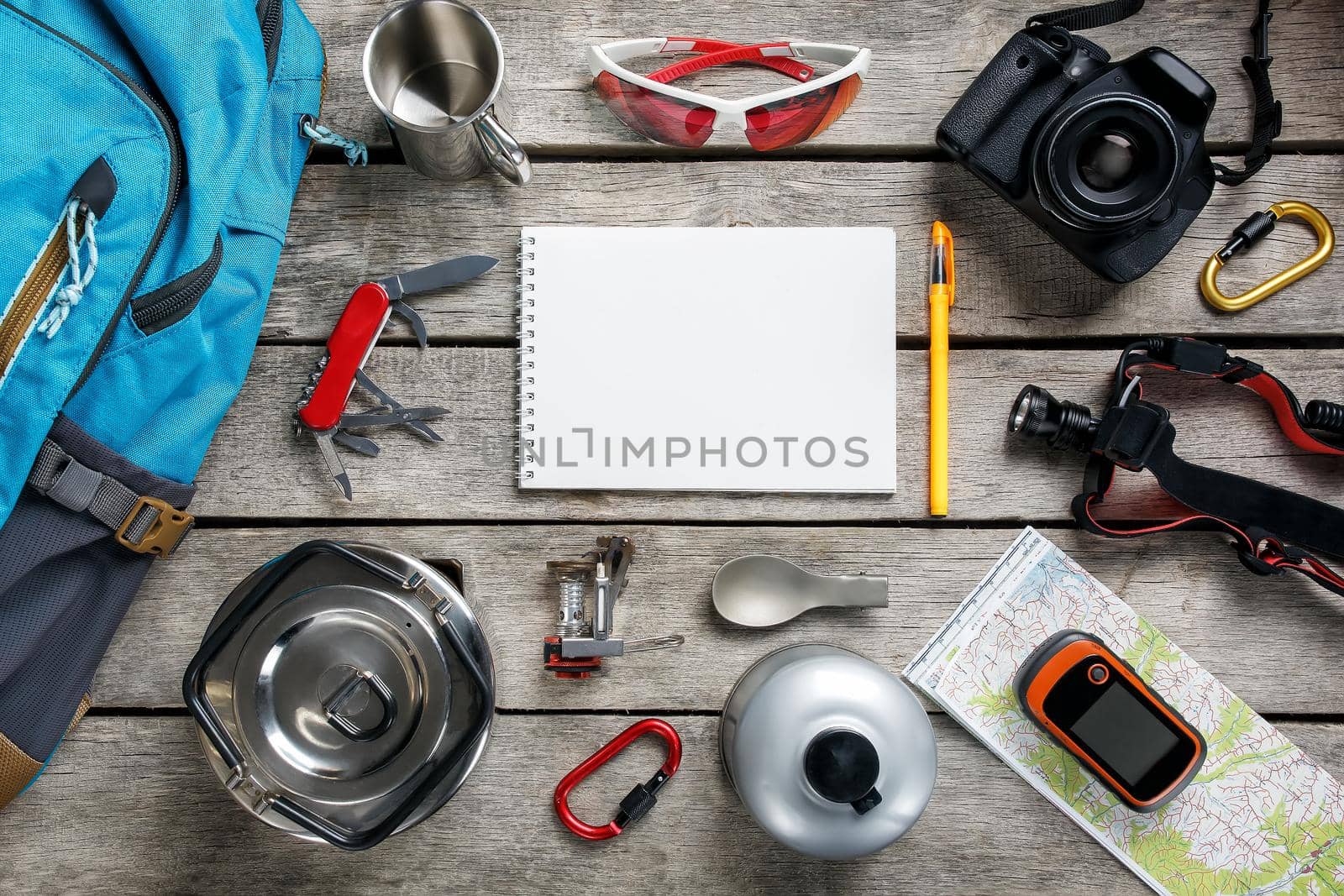 Top view of tourist equipment for travel and tourism on a rustic light wooden floor with an empty space in the middle. Items include a kettle, a card, a knife, a rope, a carbine, a flashlight, shoes, GPS, a gas bottle, a burner, a spoon, a mug, a camera, glasses, a backpack.