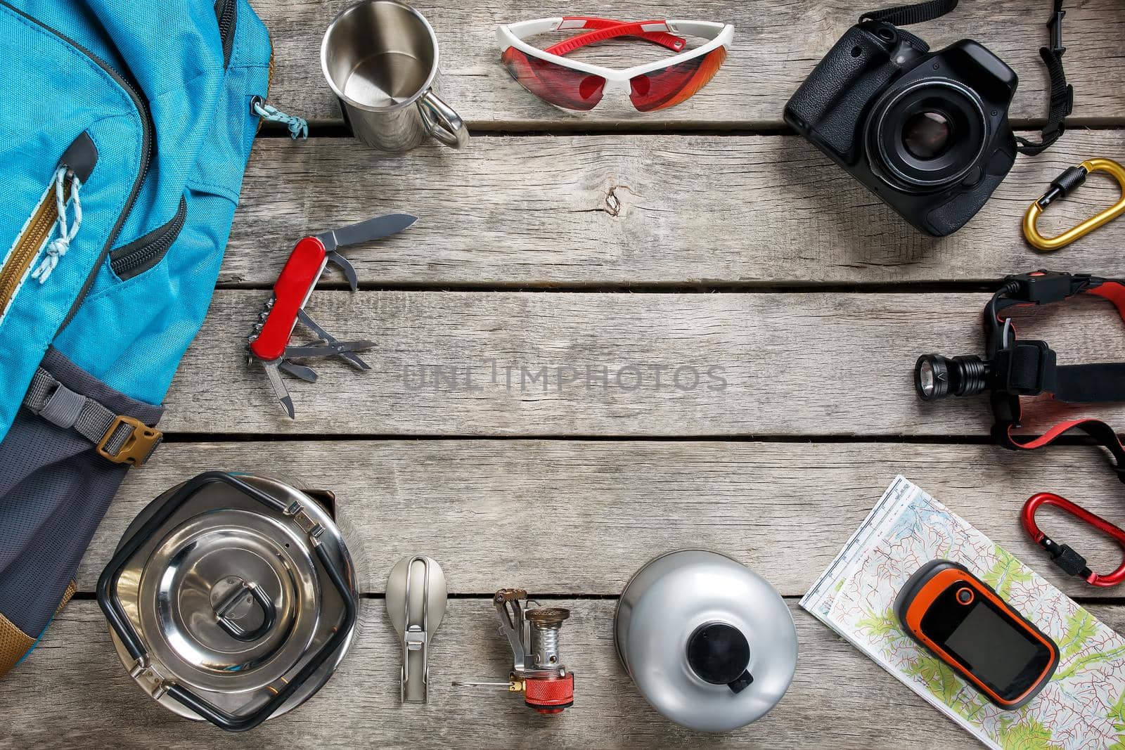 Top view of tourist equipment for travel and tourism on a rustic light wooden floor with an empty space in the middle. Items include a kettle, a card, a knife, a rope, a carbine, a flashlight, shoes, GPS, a gas bottle, a burner, a spoon, a mug, a camera, glasses, a backpack.