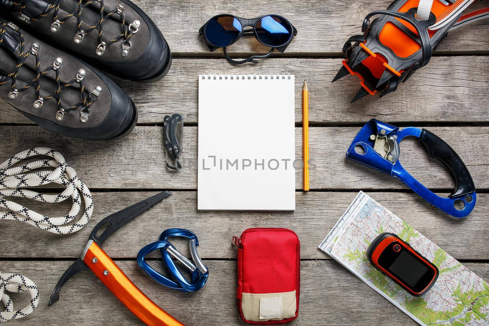 Top view of tourist equipment for a mountain trip on a rustic light wooden floor with a notebook, pencil and empty space in the middle. Items include glasses, a card, a multi-knife, a rope, a carbine, a flashlight, a first aid kit, an ice ax, a GPS, mountaineering cats.