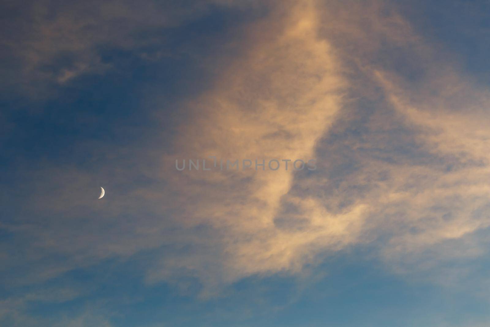 Growing moon in the blue sky among the clouds lit by the sun at sunset. Blue, pink and orange color