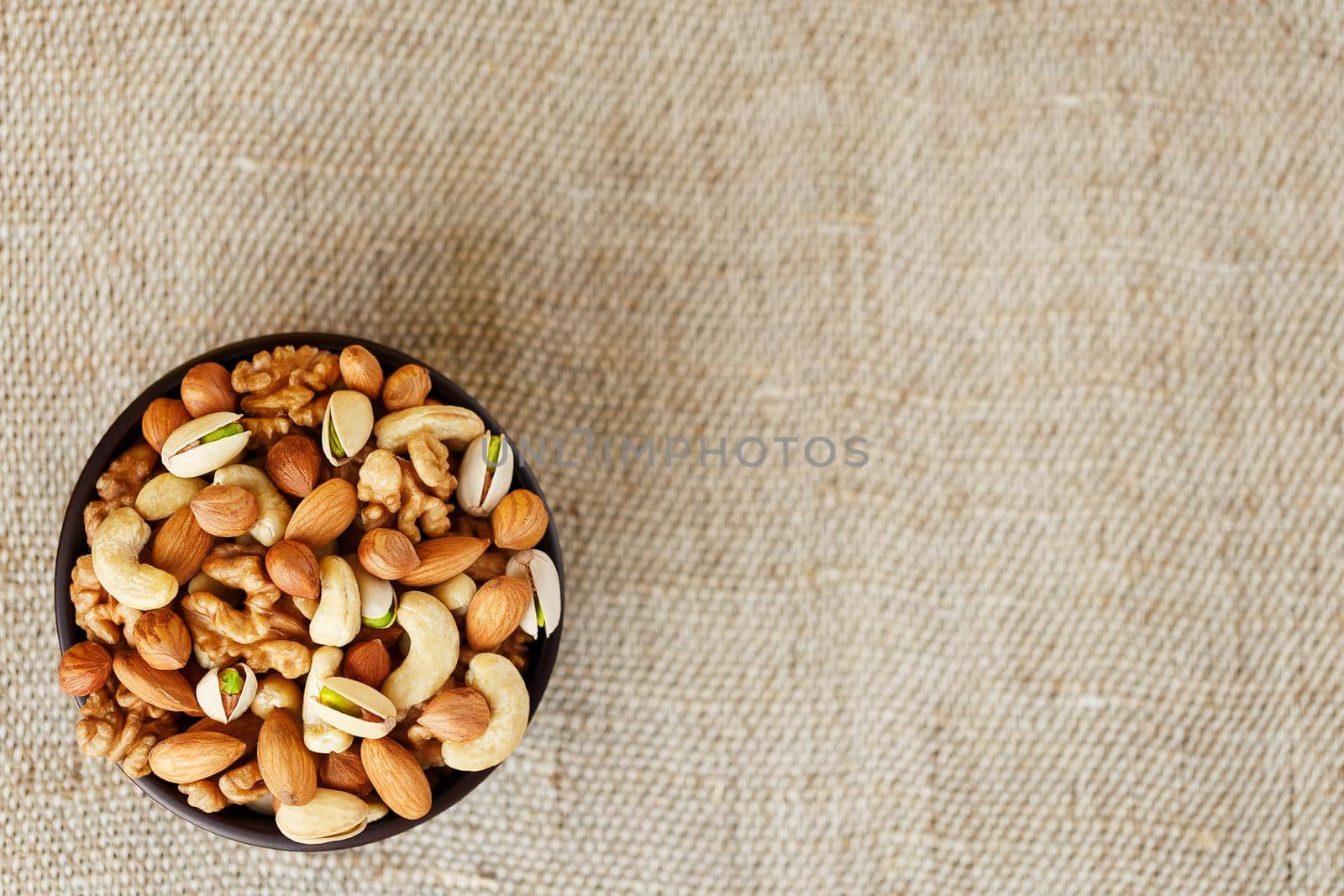 Mix of different nuts in a wooden cup against the background of fabric from burlap. Nuts as structure and background, macro. Top view. by AlexGrec