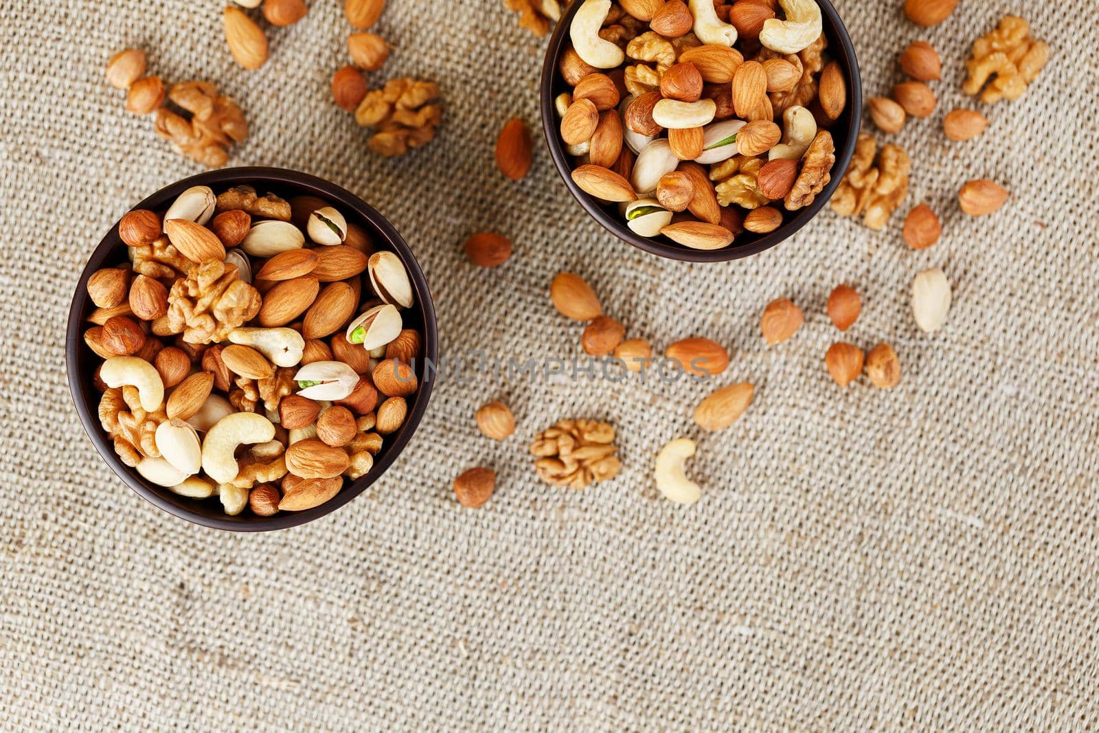 A mixture of cashew nuts, almond nuts, pistachios, hazelnuts and walnuts in a wooden cup against the background of burlap fabric. Nuts as structure and background, macro. Two cups of nuts.