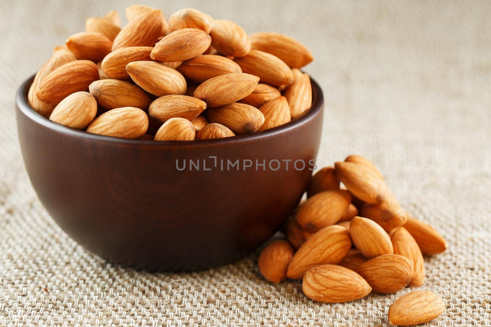 Almonds in a wooden cup on a burlap cloth background. Golden almond closeup in dark brown cup