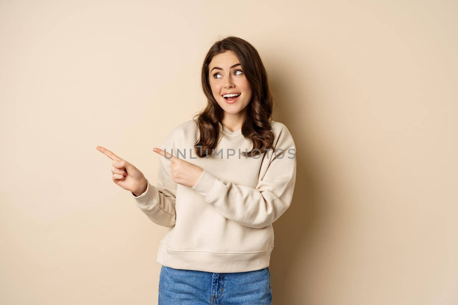 Cheerful attractive female model pointing fingers left, showing store advertisement, banner or logo, standing in sweater over beige background by Benzoix