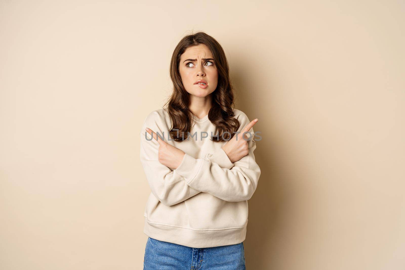 Complicated and indecisive woman pointing sideways, cant make choice, struggle with decision, choosing between two options, standing over beige background.