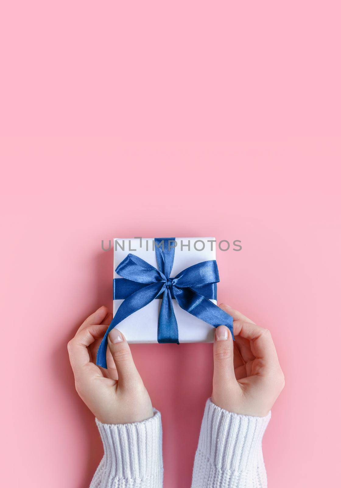 Female Hands in sweater holding gift in white wrapping paper on pink background. St. Valentines Day, love, tenderness, friendship, Birthday, Christmas, care concept. Cozy, festive, romantic wallpaper