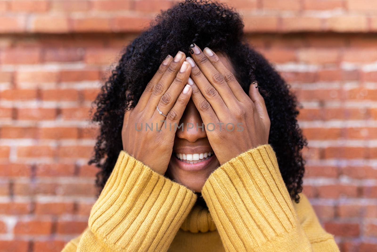 Smiling African American woman with curly hair covering her eyes with hands. by Hoverstock