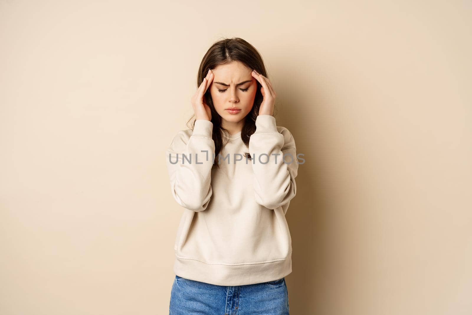 Woman with headache massaging her head temples and feeling sick, has migraine, standing over beige background.
