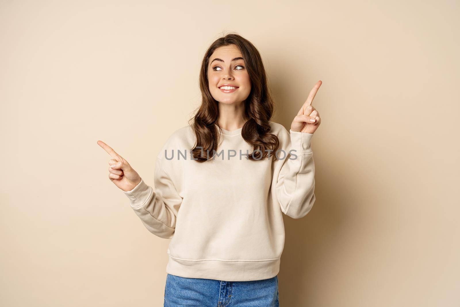 Cheerful caucasian woman showing directions, two ways, pointing sideways at variants, choices for customer, standing over beige background.