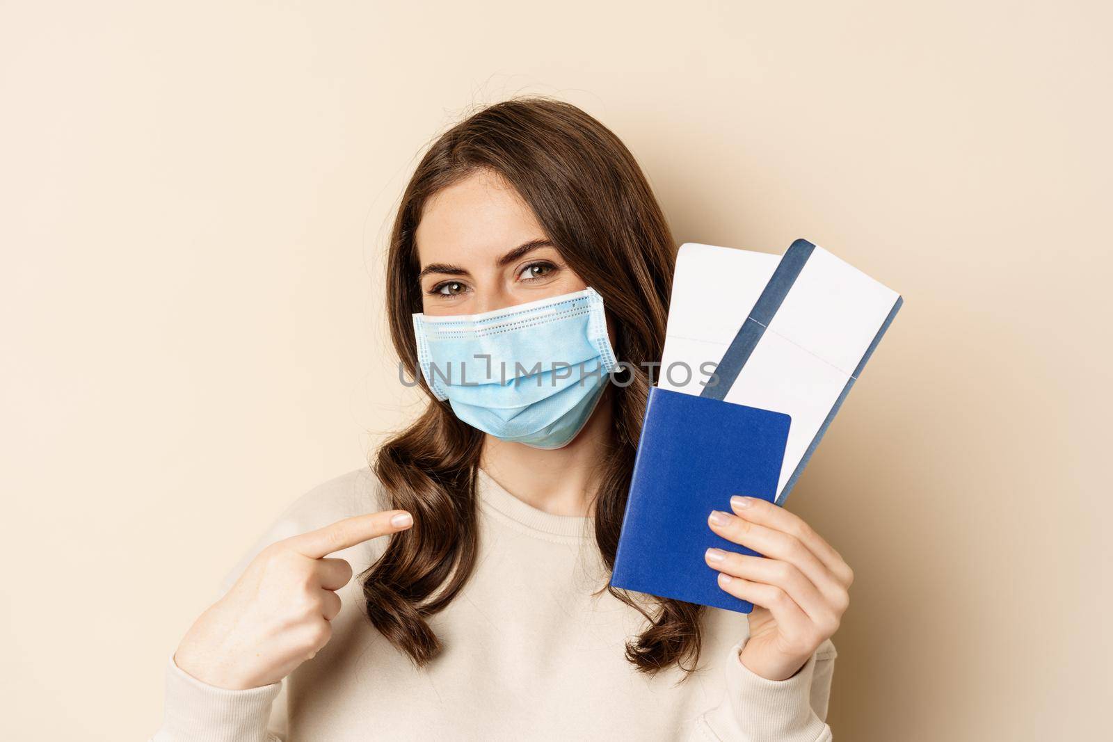 Travel and covid-19 pandemic. Girl in medical face mask travelling, showing passport with two tickets on plane, standing against beige background.