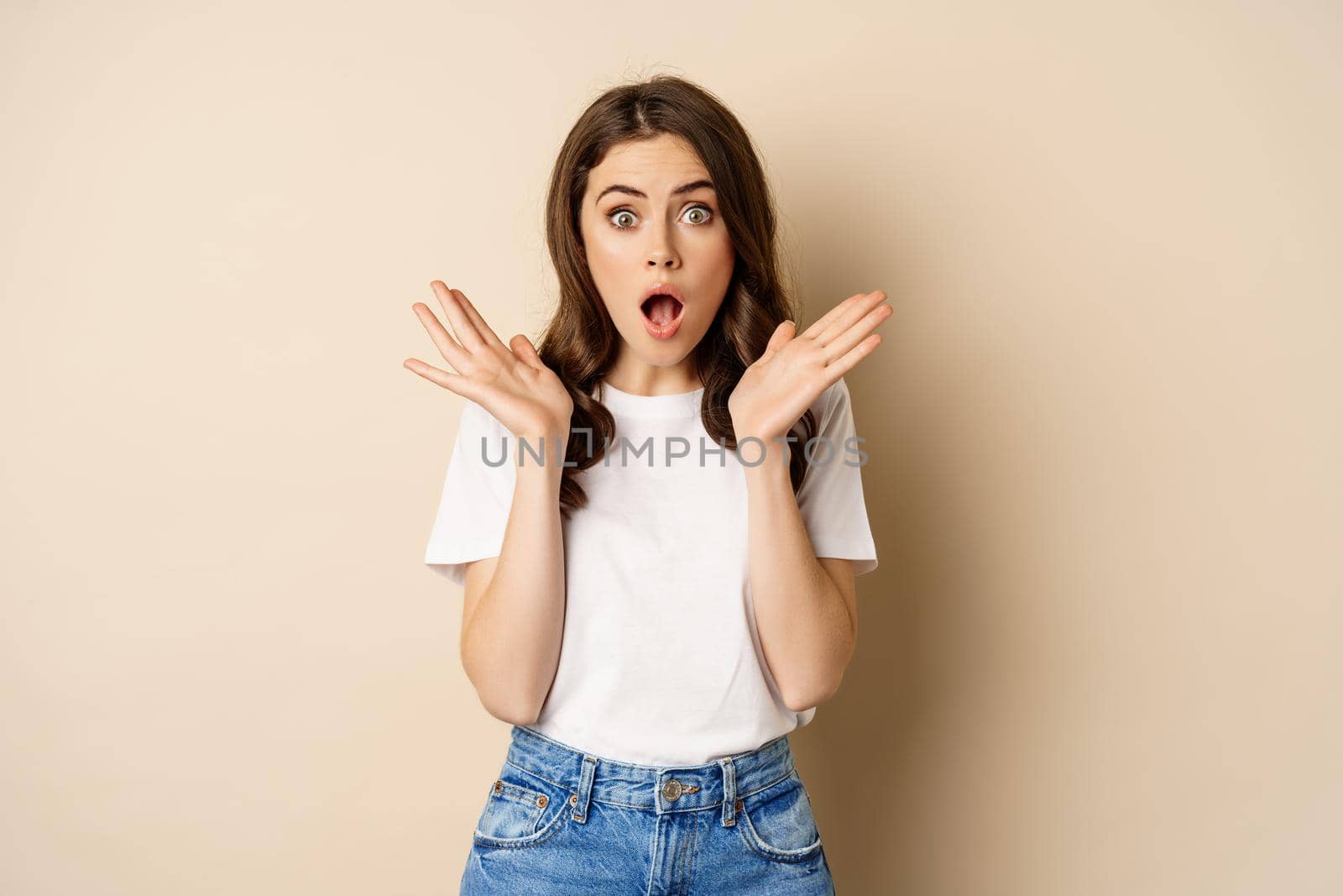 Portrait of stylish young woman gasping, looking surprised and amazed, impressed by smth, standing in t-shirt and jeans over beige background.