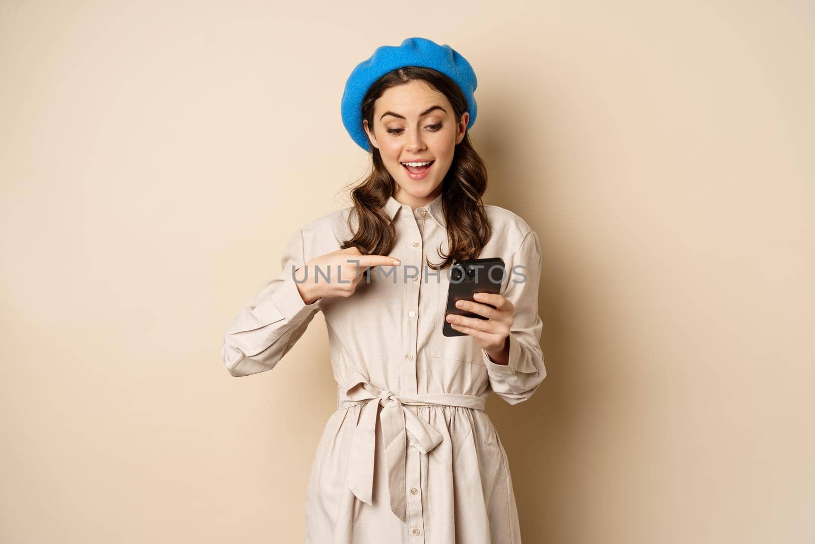 Portrait of stylish modern woman in outerwear, pointing at mobile phone screen and looking happy, smiling pleased, standing over beige background.