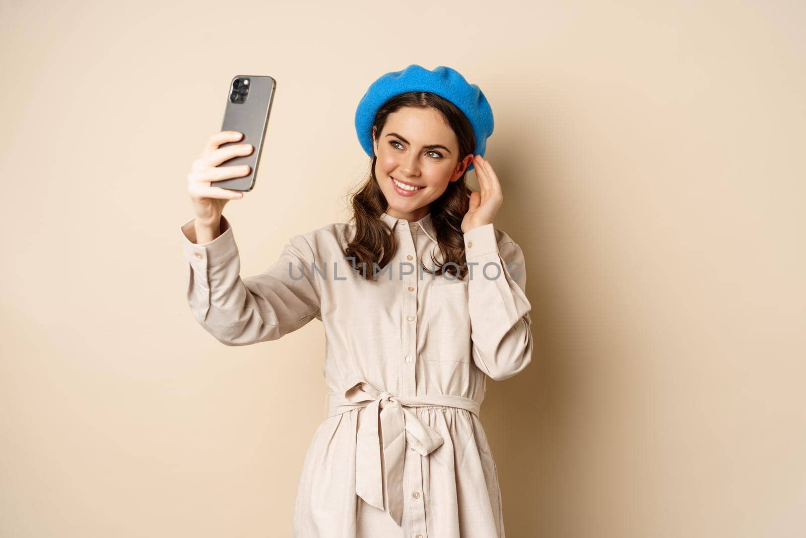 Beautiful girl taking selfie on smartphone in coat and cute hat, smiling for photo and looking happy, standing over beige background by Benzoix