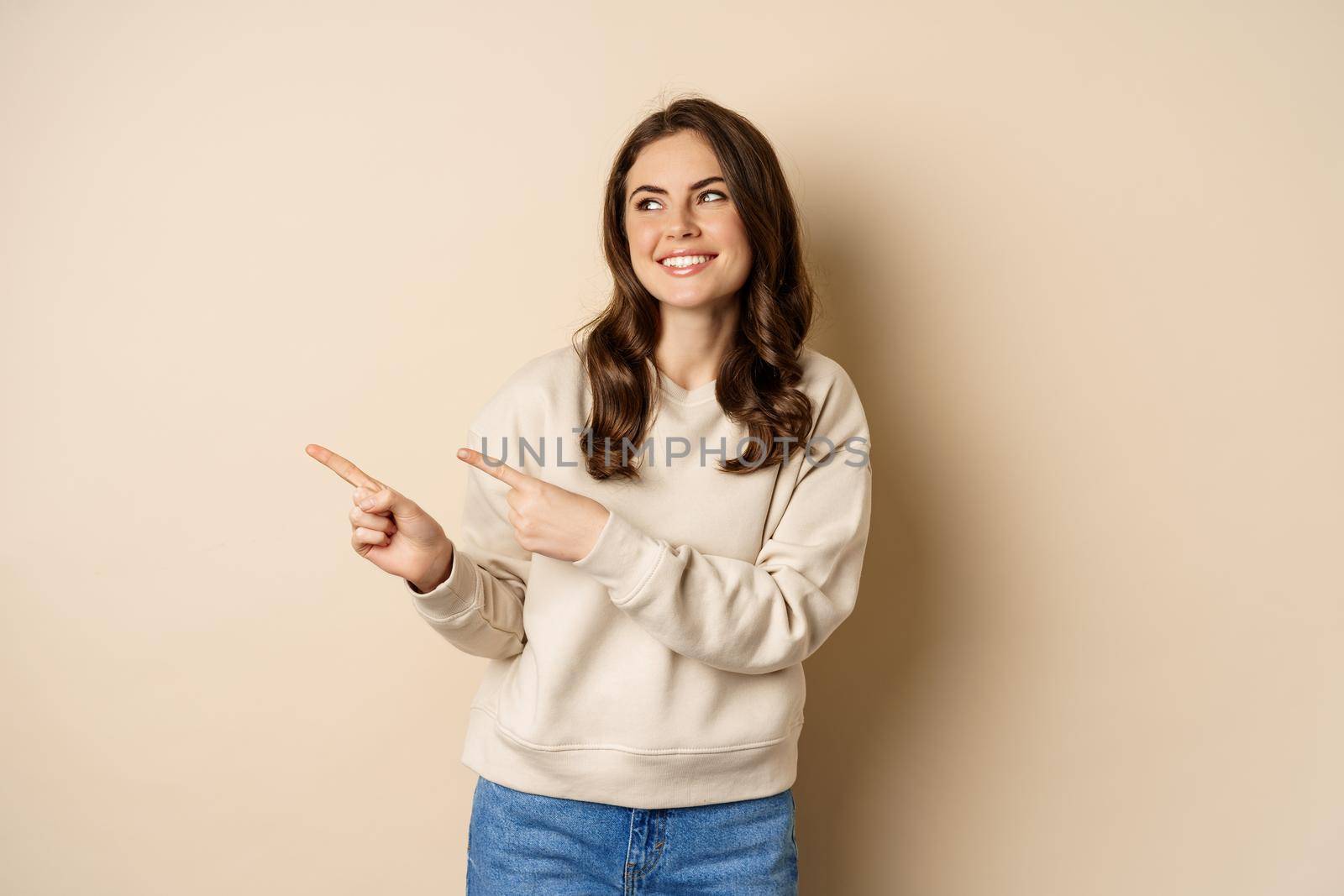Beautiful girl pointing and looking left, laughing and smiling happy while showing advertisement, logo or store banner, beige background.