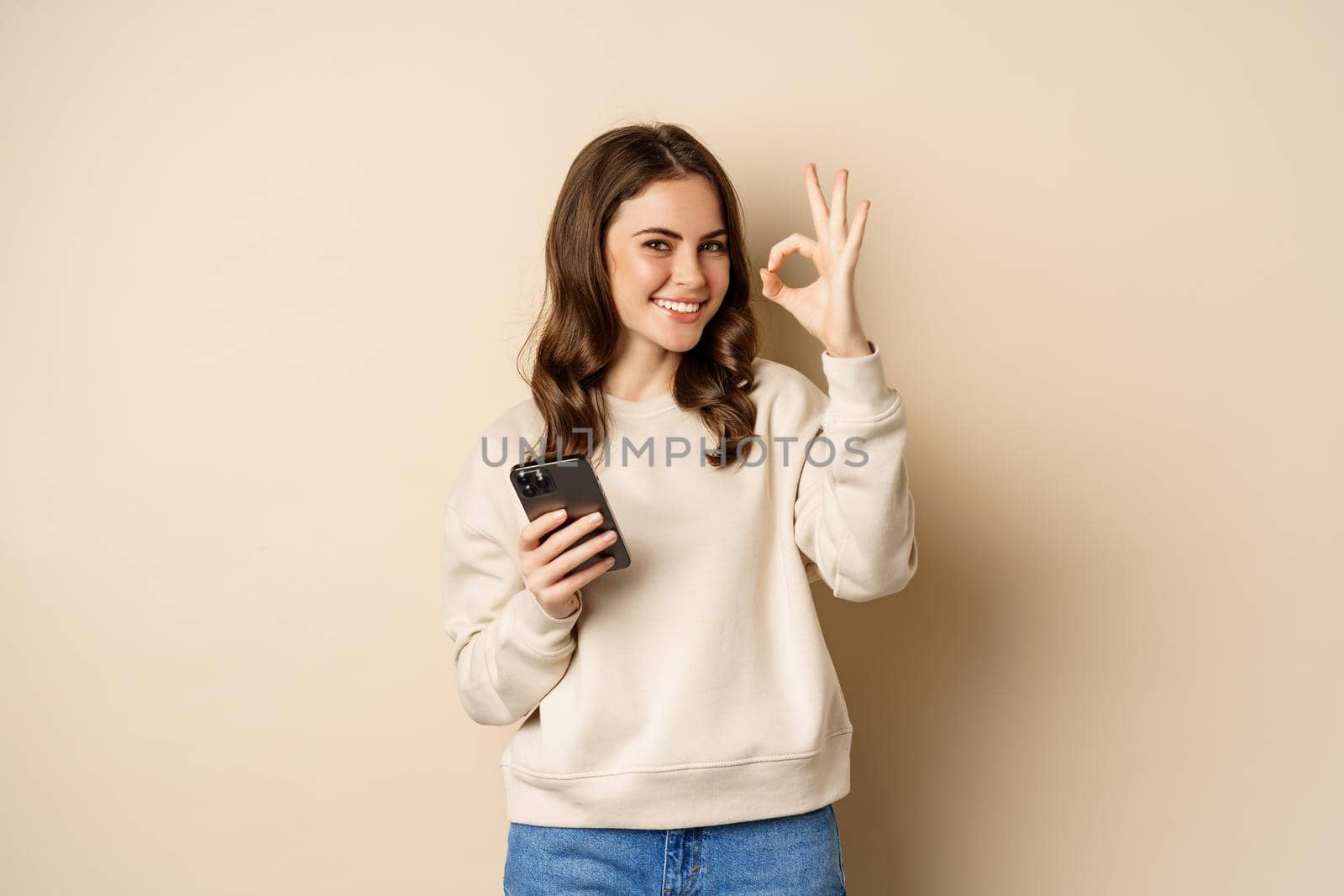 Beautiful woman holding cellphone, mobile phone and okay sign, recommending application, shopping app, standing over beige background by Benzoix