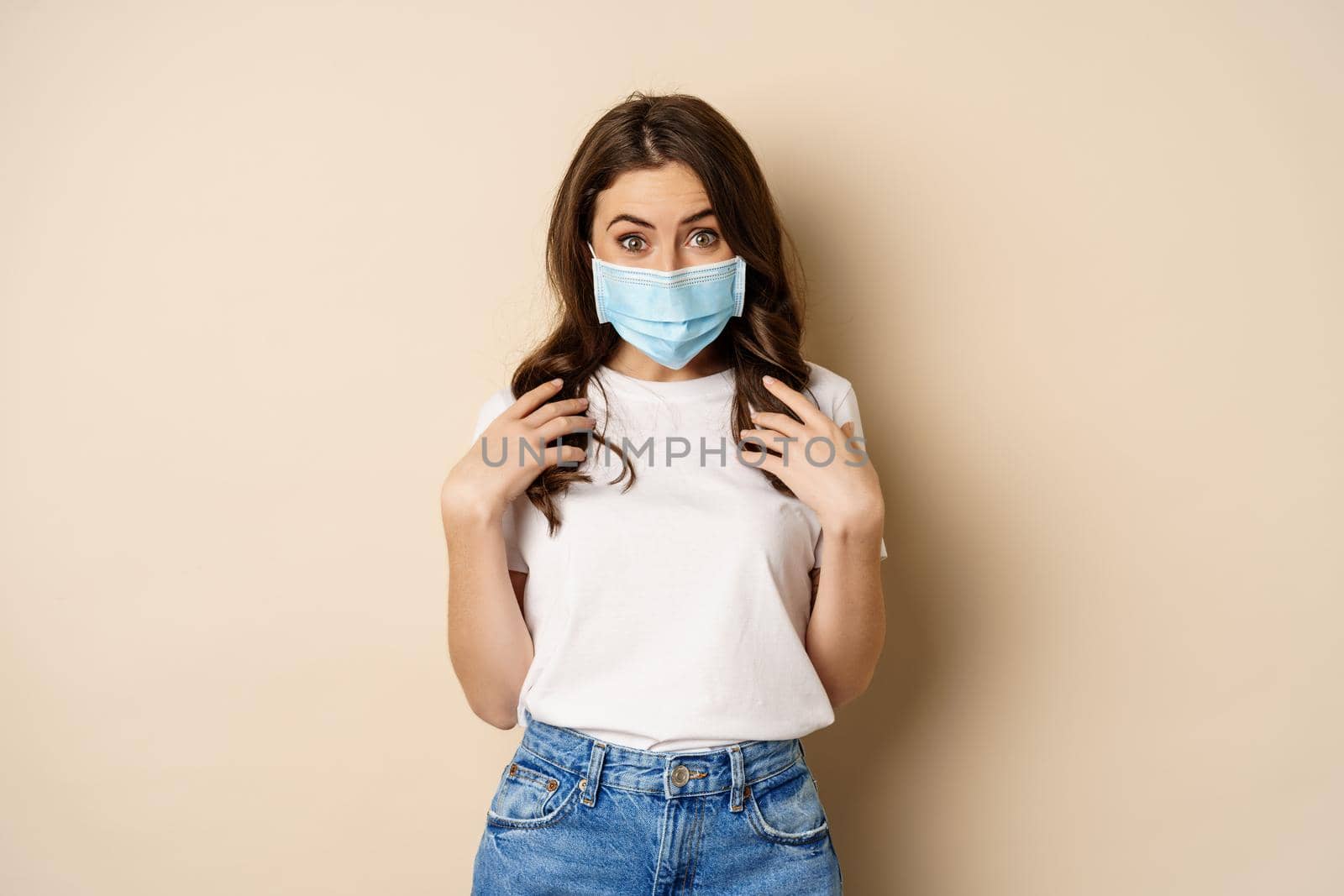 Covid-19 and people concept. Young woman in medical face mask and t-shirt, looking surprised at camera, eyes in disbelief, standing over beige background by Benzoix