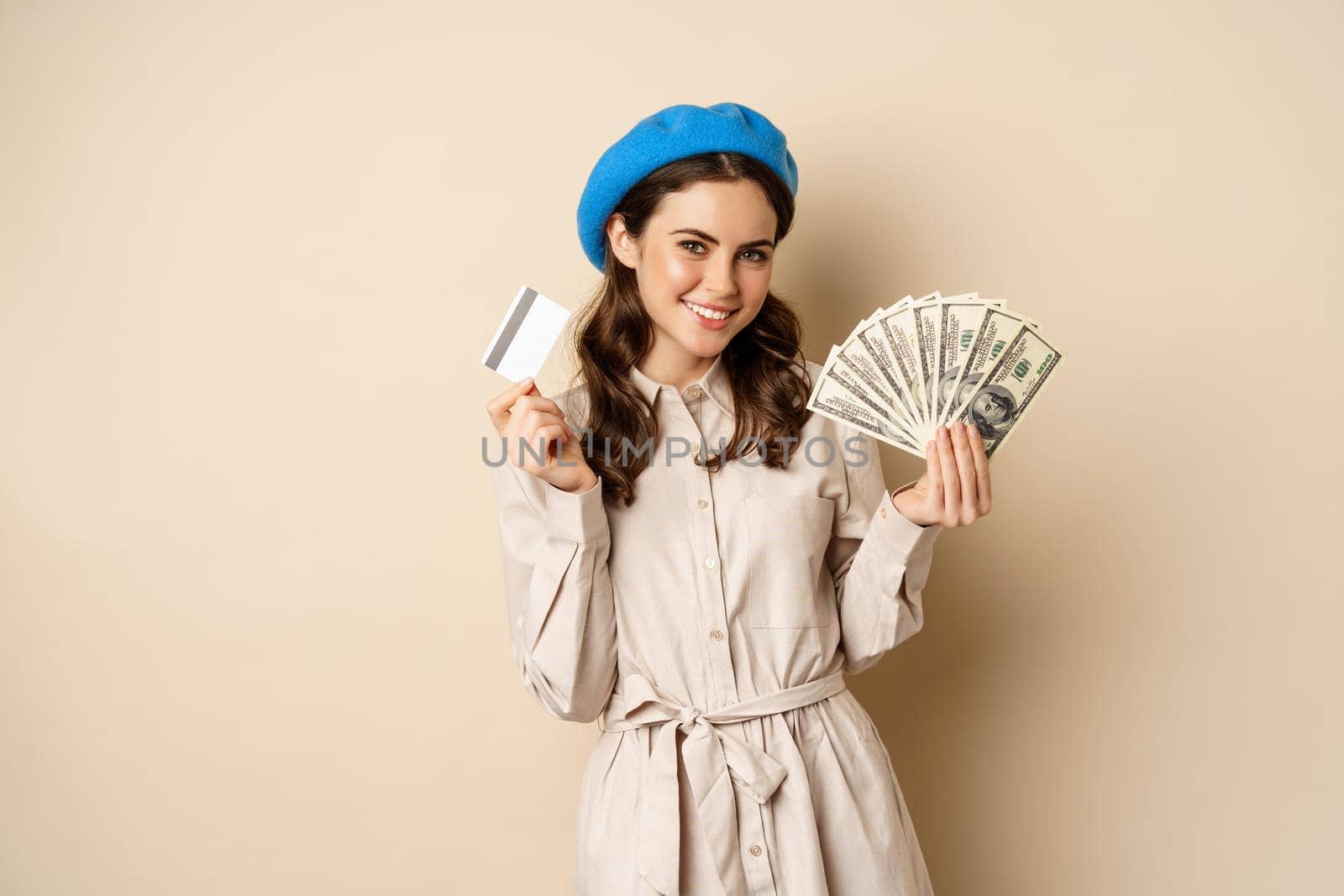 Microcredit and money concept. Young stylish woman showing credit card and dollars cash, smiling happy and satisfied, standing over beige background by Benzoix