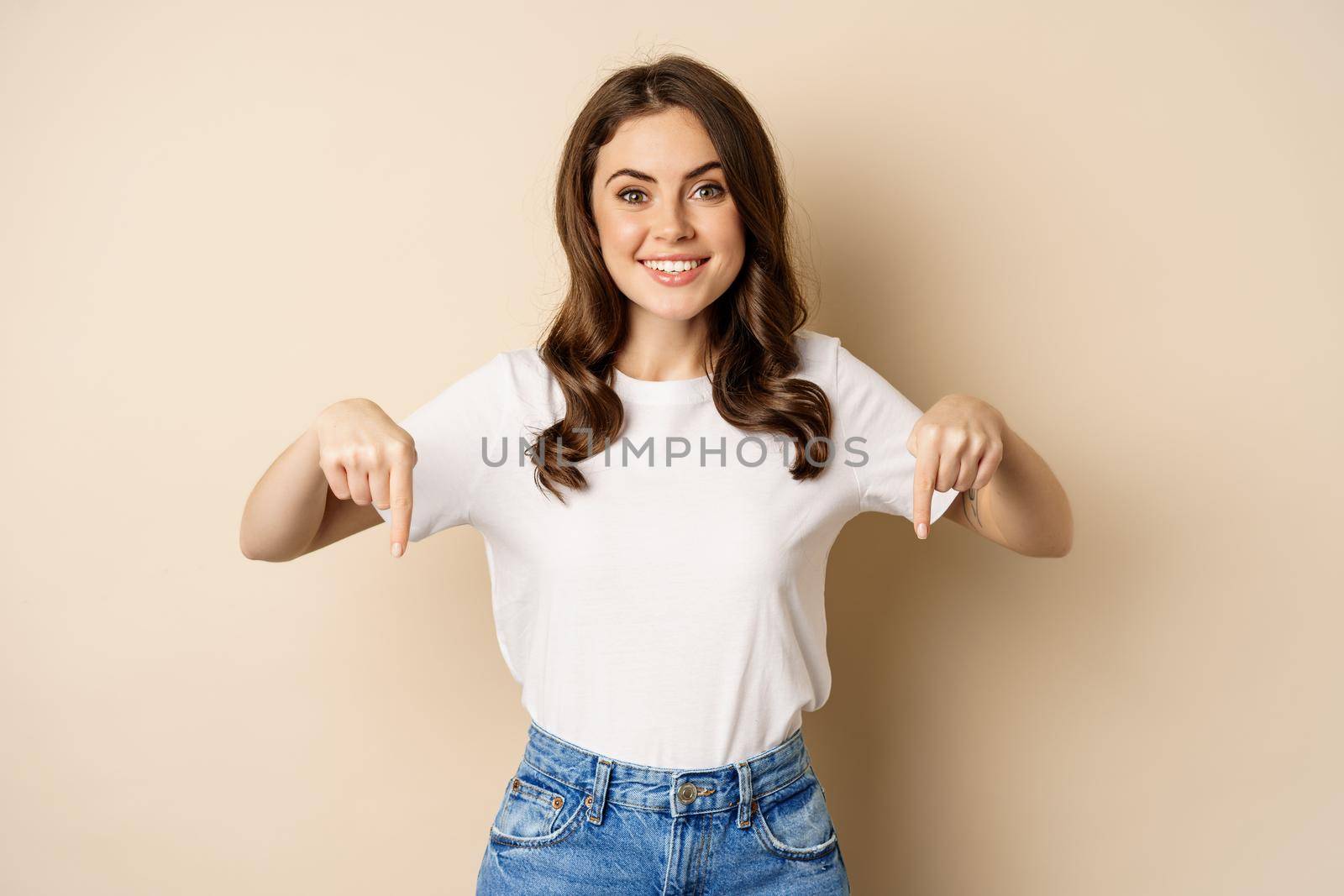 Smiling happy girl pointing fingers down and showing banner, company logo below, click link, standing over beige background.