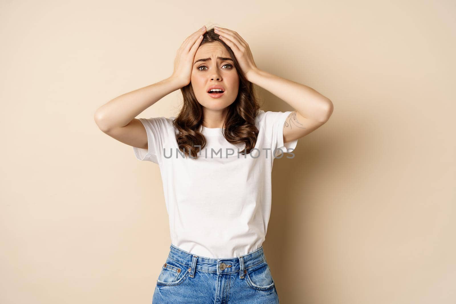 Young woman facing disaster, looking anxious in panic, holding hands on head frustrated, standing over beige background.