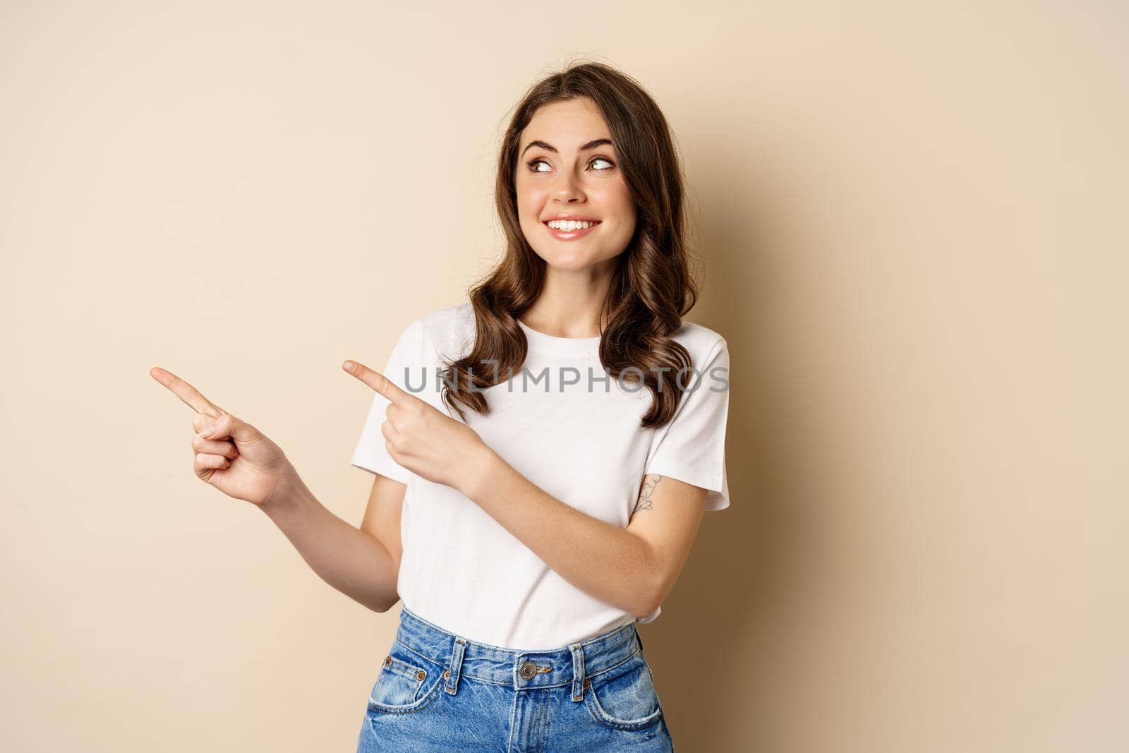 Stylish young caucasian woman smiling, pointing fingers left, showing advertisement, promo offer, standing against beige background.
