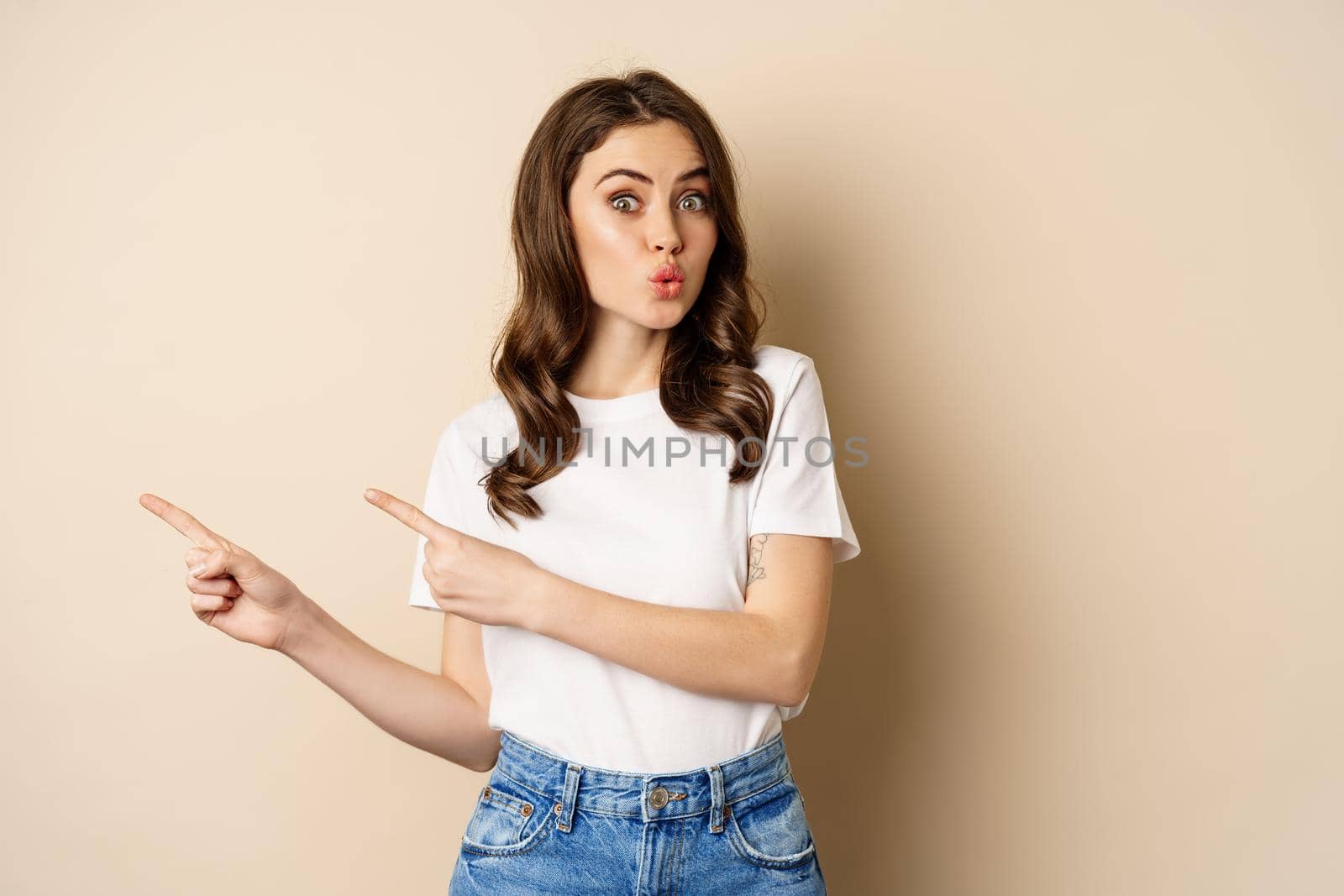 Stylish young caucasian woman smiling, pointing fingers left, showing advertisement, promo offer, standing against beige background by Benzoix