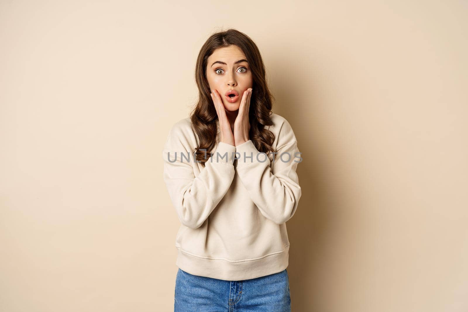 Image of surprised, amazed brunette girl gasping, looking fascinated at camera, standing over beige background.