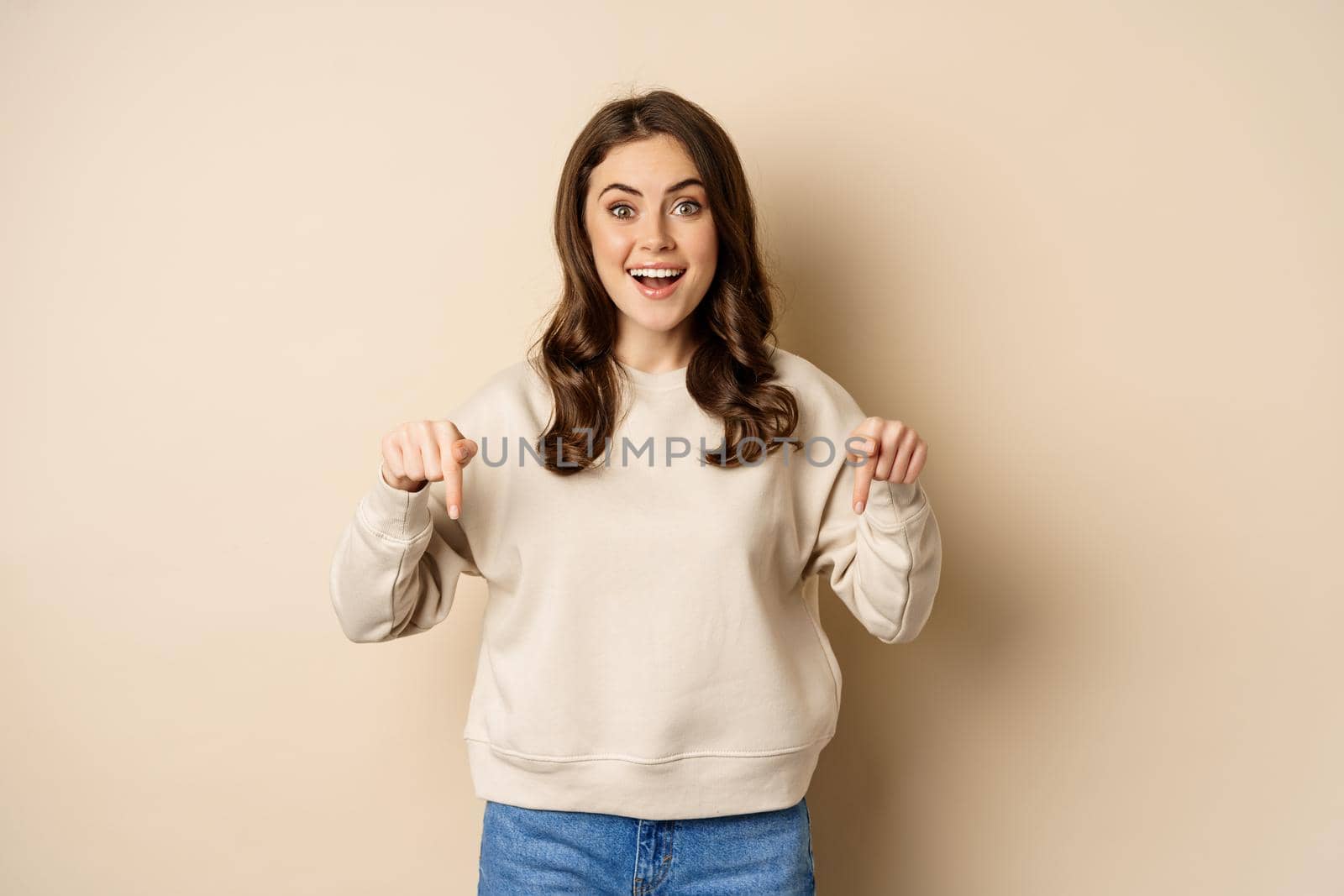 Enthusiastic attractive girl pointing fingers down, showing advertisement, link or promo below, standing over beige background by Benzoix
