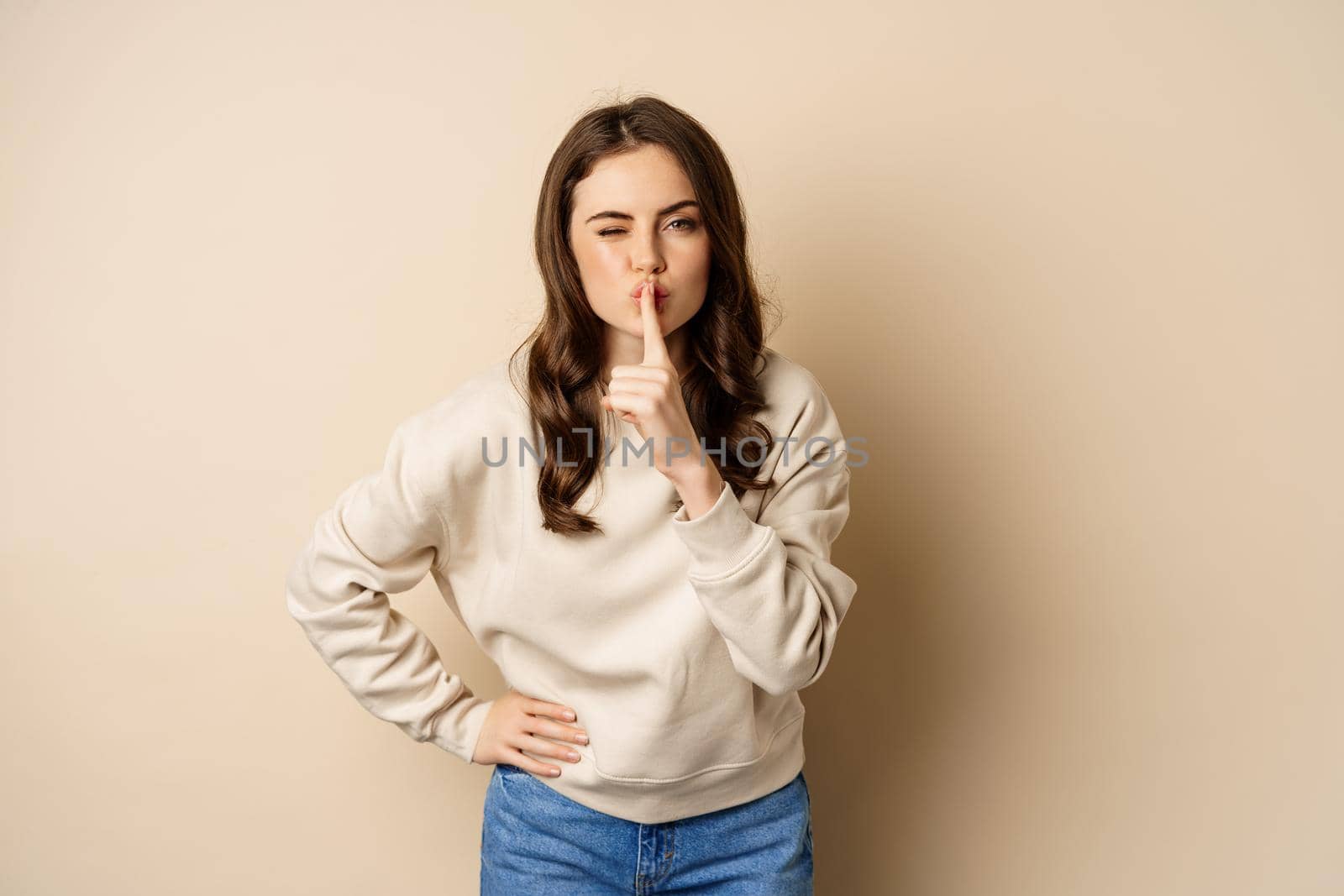 Happy coquettish girl say shh, shushing, keep secret, be quiet gesture, smiling at camera, standing over beige background.