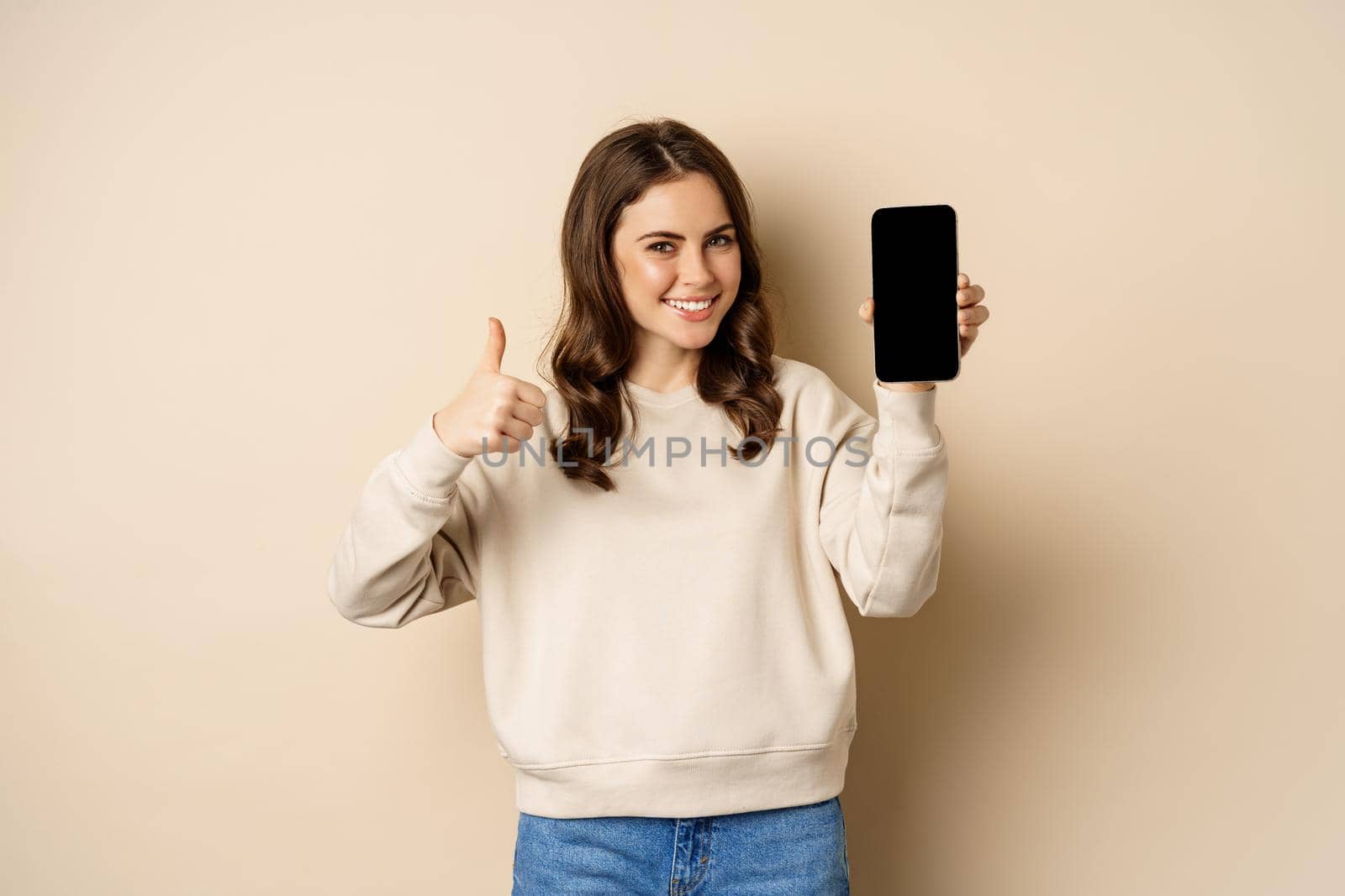 Attractive smiling woman showing thumbs up and smartphone screen, standing over beige background.