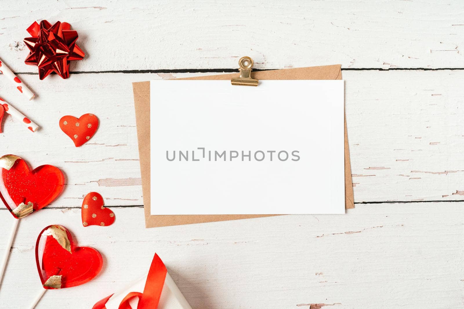 Flat lay of paper envelope with blank mockup greeting paper card. White wooden table background with Valentine day gift, letter, heart shape, lollipops, confetti, decor. Top view, mock up invitations