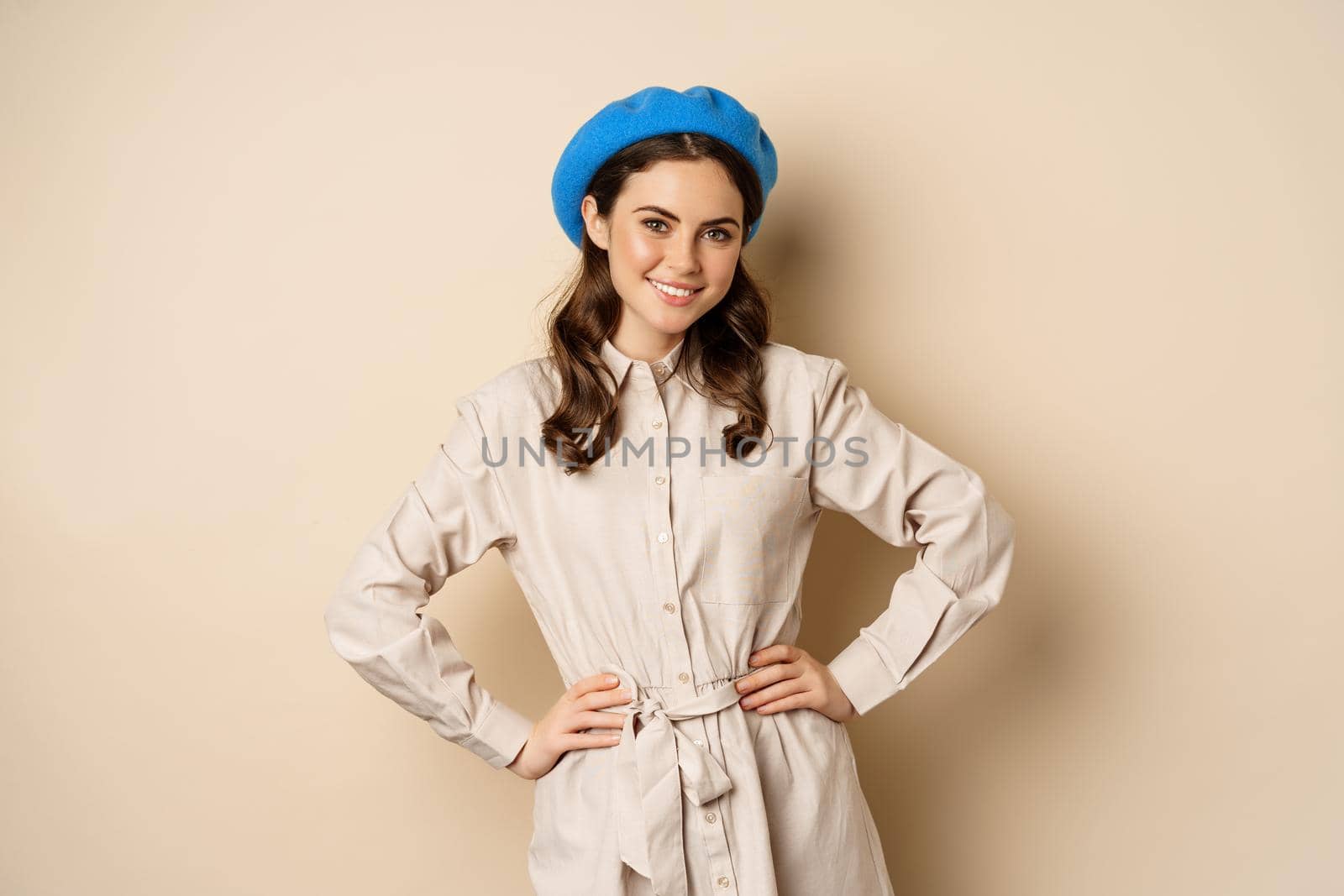 Portrait of trendy woman in trench coat and french hat, smiling and looking happy, posing in stylish outfit against beige background.