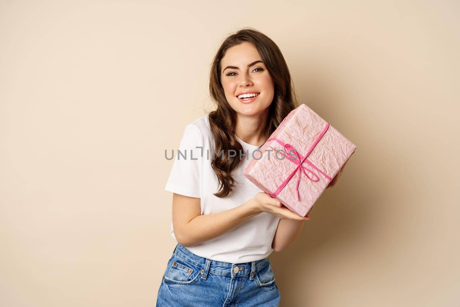 Celebration and holidays concept. Happy young woman holding gift wrapped in pink box, receive present, looking amazed and surprised, standing over beige background.