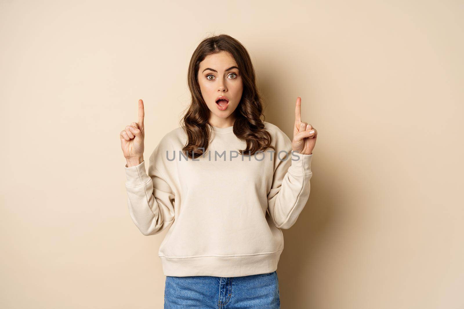 Surprised beautiful girl showing advertisement, pointing fingers on top, store sale or website link up, standing over beige background.
