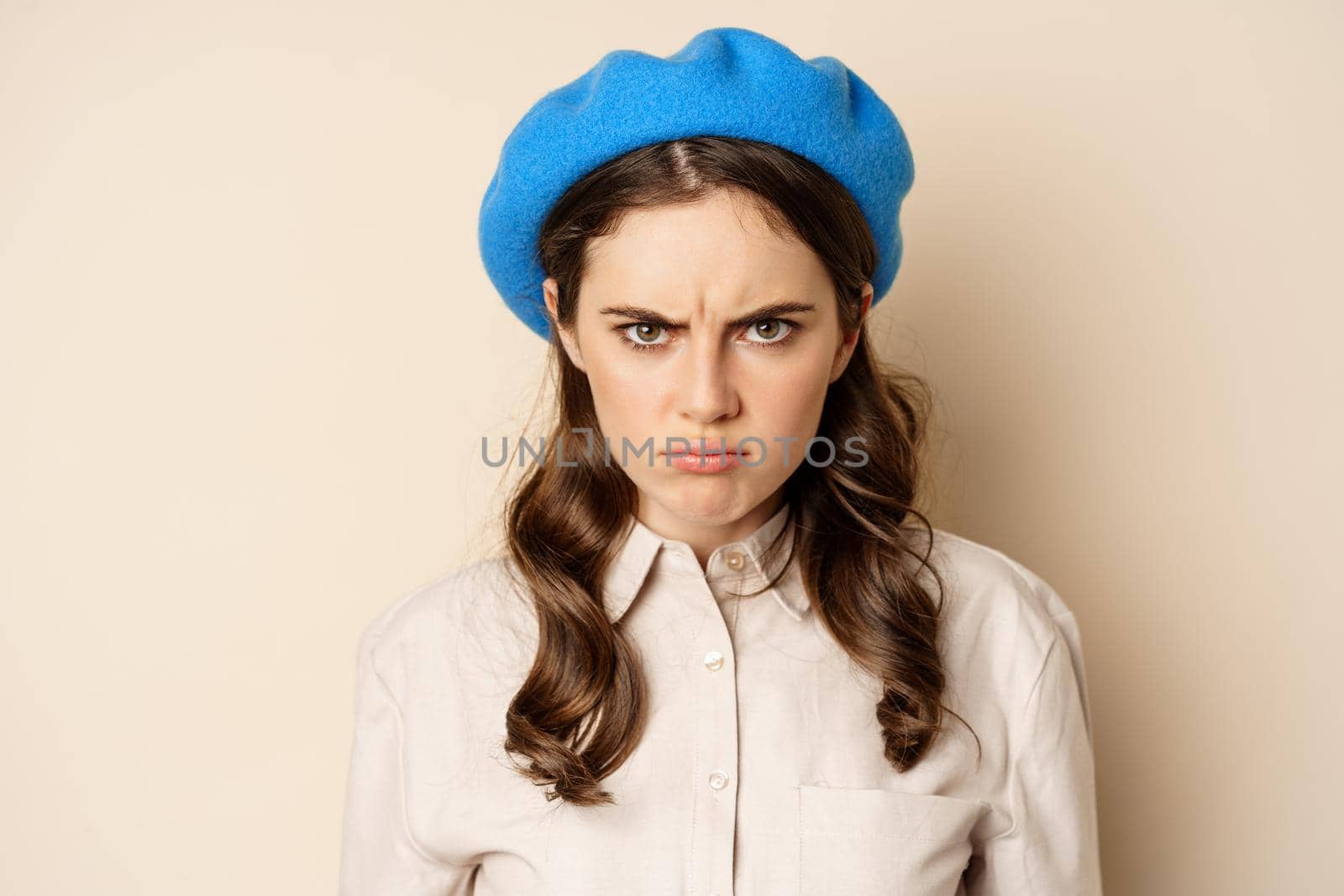 Close up portrait of young beautiful woman sulking, looking angry and disappointed, grimacing displeased, standing over beige background by Benzoix