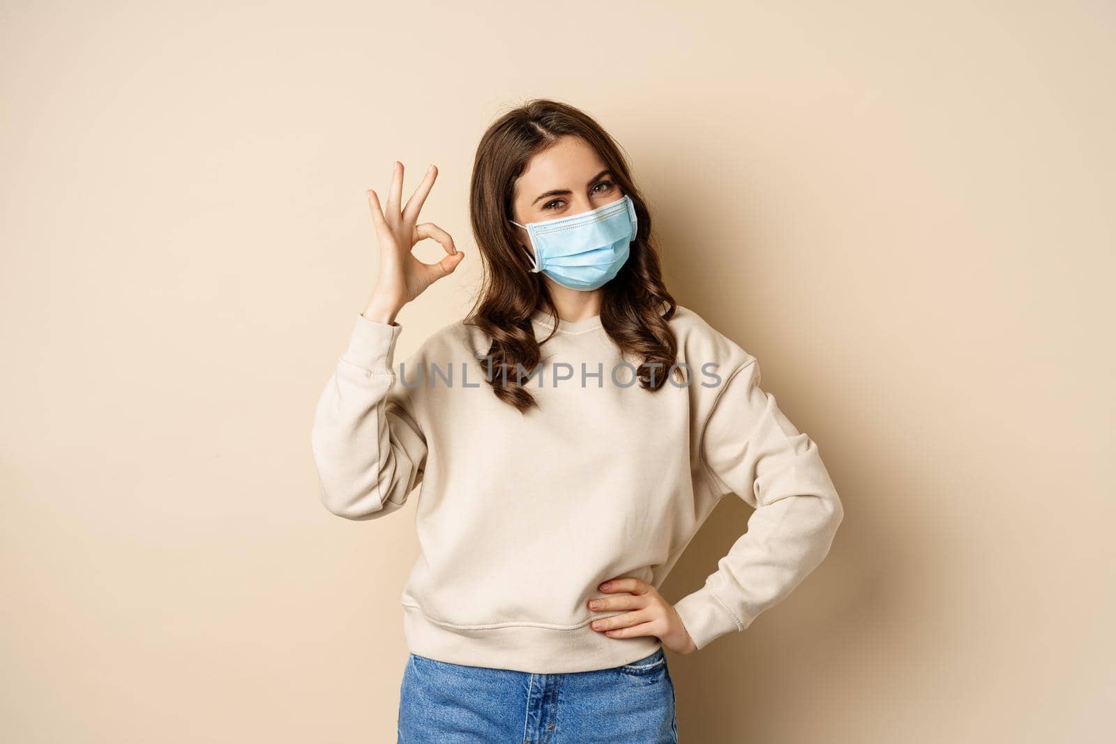 Covid-19, pandemic and quarantine concept. Young woman wears medical face mask during coronavirus omicron outbreak, showing okay sign, standing over beige background.