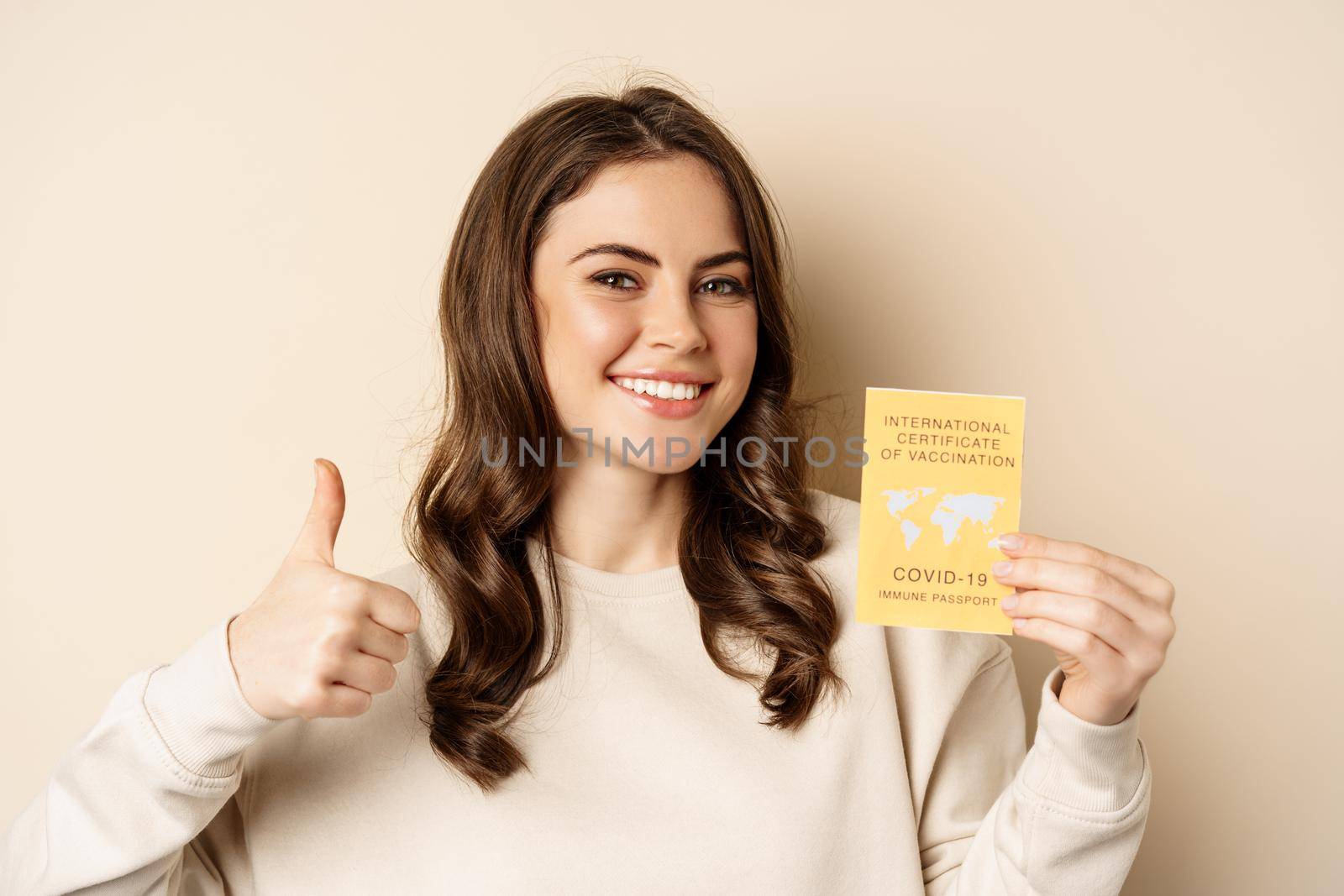 Covid-19 vaccination concept. Portrait of happy healthy girl showing coronavirus international vaccination certificate and thumb up, travelling during pandemic, beige background by Benzoix