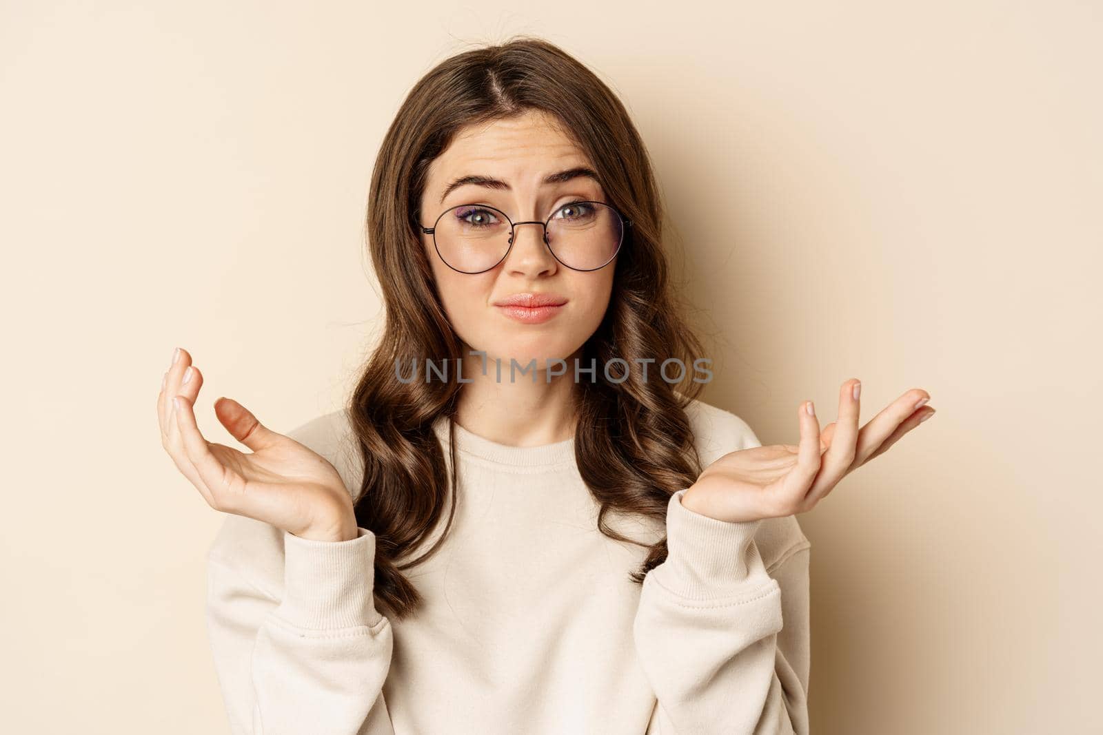 Young indecisive woman in glasses shrugging shoulders and looking confused, dont know, cant understand, standing over beige background.