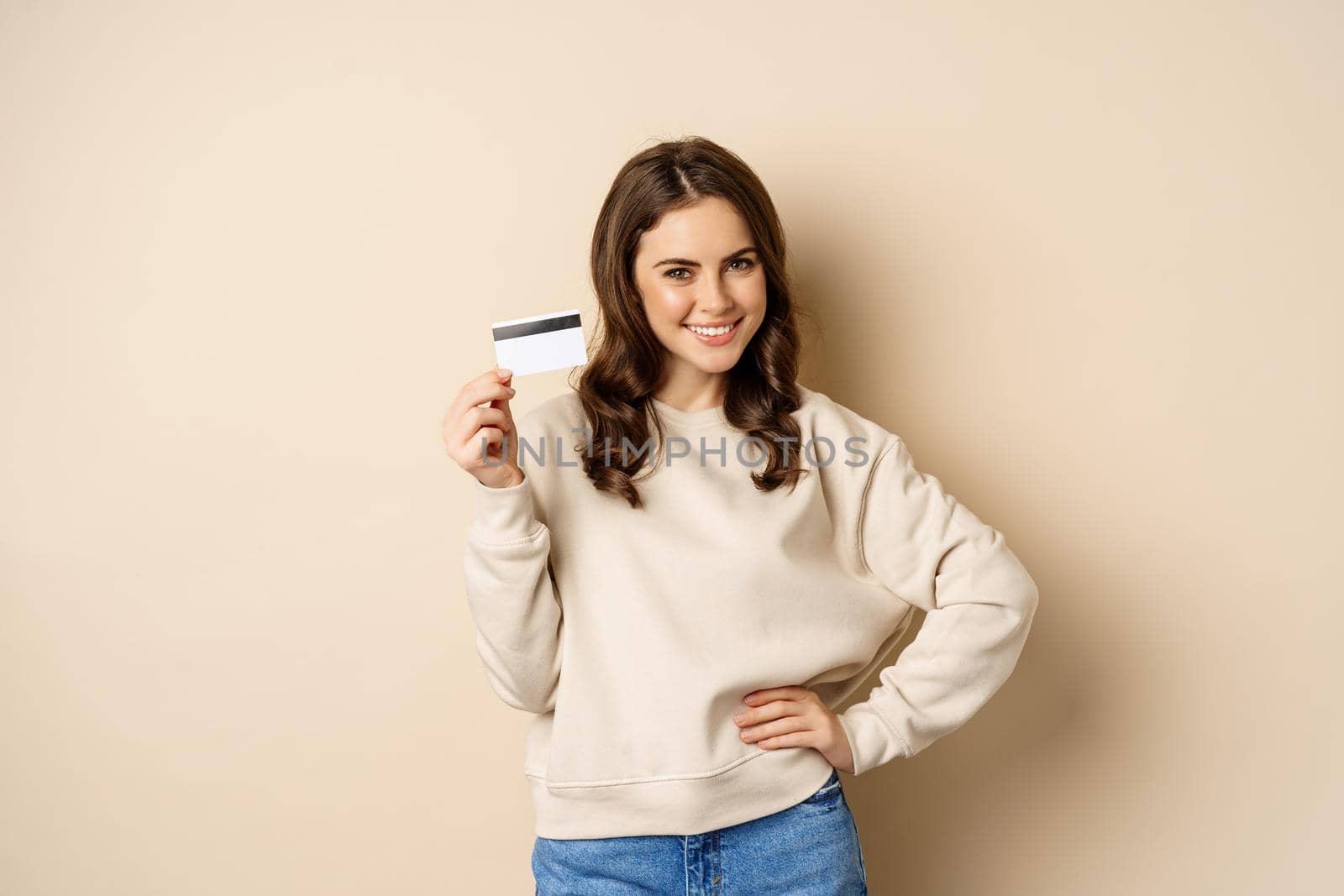 Smiling beautiful woman showing credit card, concept of shopping, contactless payment, standing over beige background by Benzoix