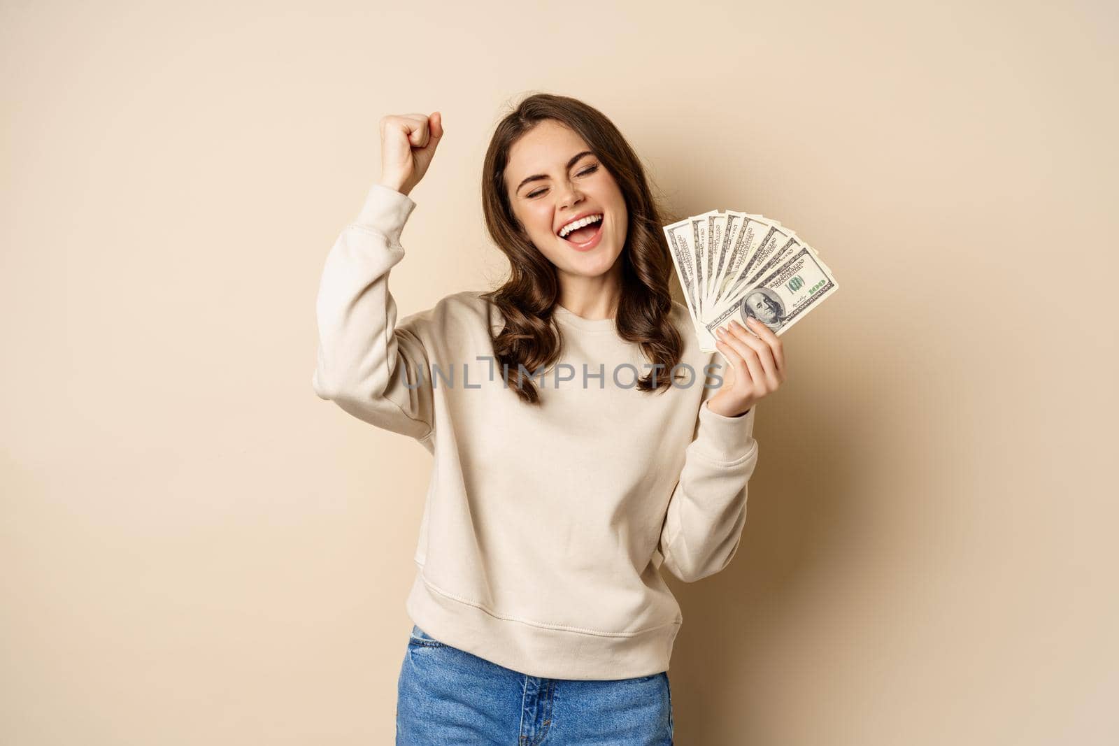 Enthusiastic modern woman winning money, got cash, celebrating and shouting of joy, standing against beige background by Benzoix