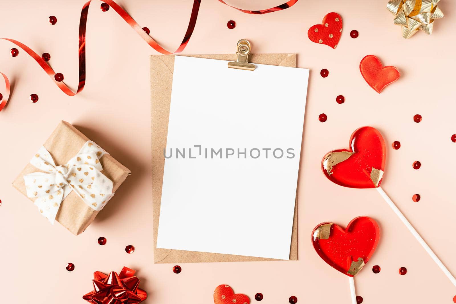 Flat lay of paper envelope with blank mockup greeting paper card. Pink table background with Valentine day gift, letter, heart shape, confetti, lollipops and decoration. Top view, mock up invitations