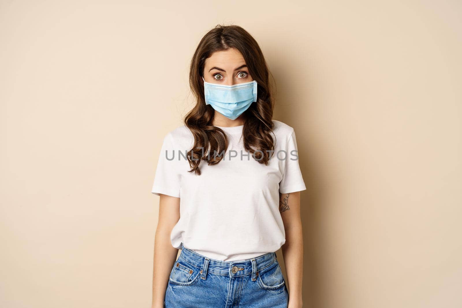 Covid-19 and people concept. Young woman in medical face mask and t-shirt, looking surprised at camera, eyes in disbelief, standing over beige background.