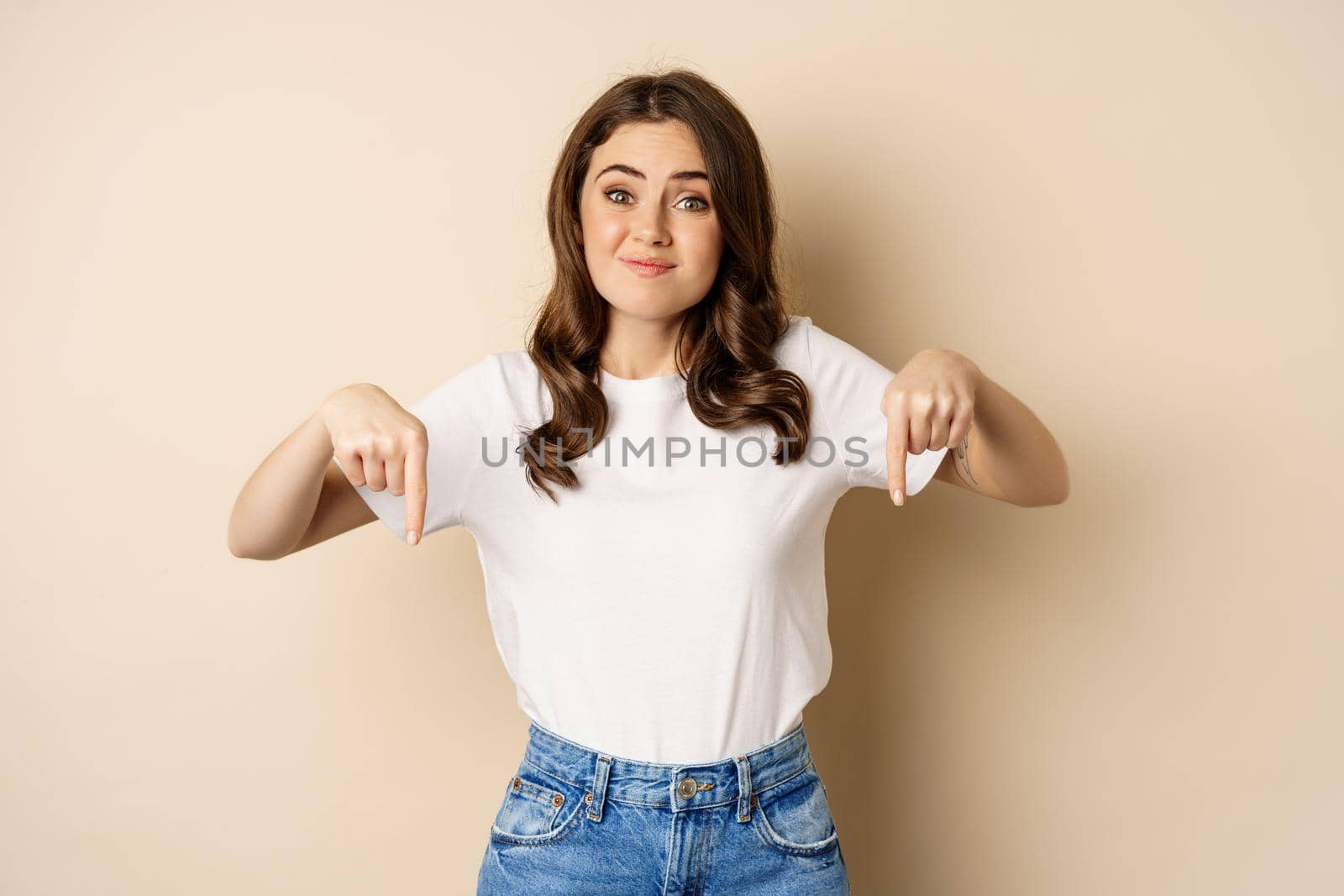 Doubtful girl pointing fingers down and smirking, looking uncomfortable while complaining, dislike smth, standing over beige background.