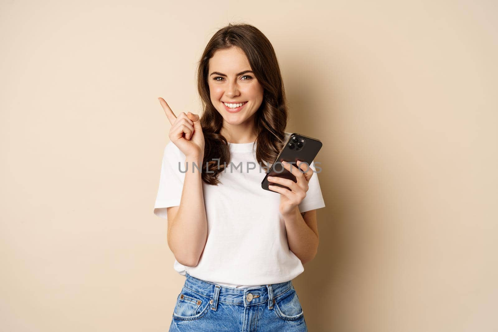 Stylish young modern woman holding mobile phone, pointing finger left and smiling, concept of shopping, beige background.