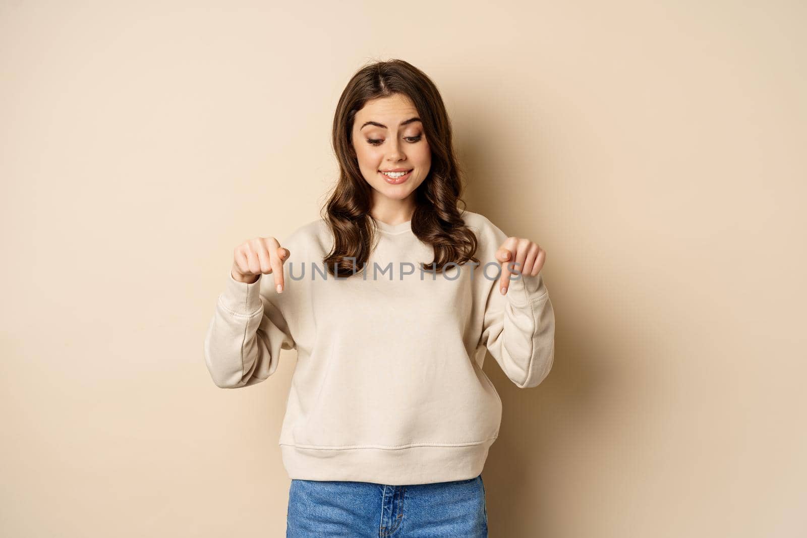 Excited adult woman pointing fingers down and smiling, looking at banner below, showing advertisement, found smth, standing over beige background by Benzoix