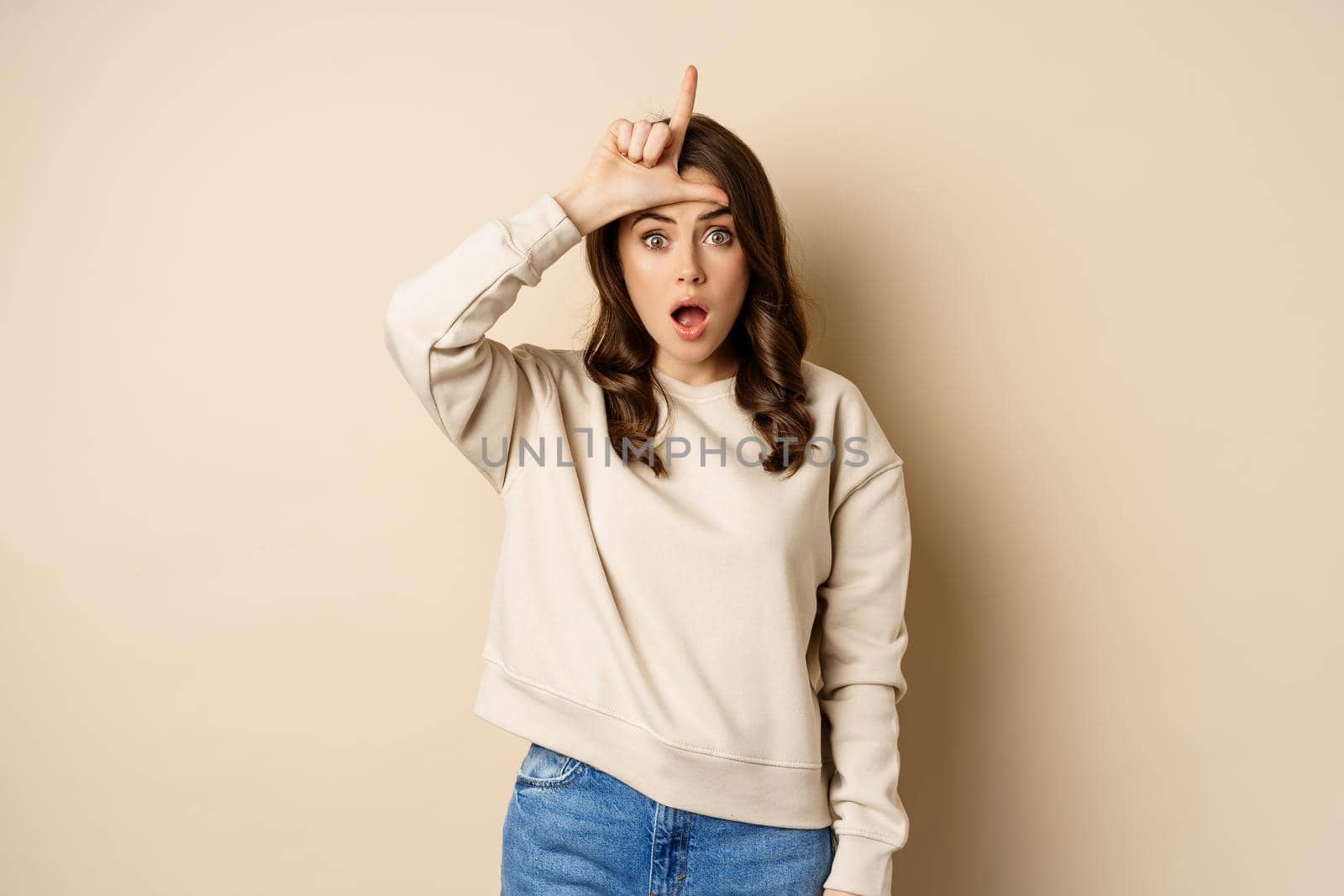 Woman showing loser sign on forehead, L word, standing over beige background. Copy space