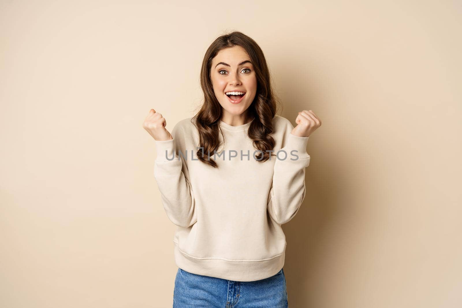 Enthusiastic brunette woman laughing and smiling, cheering, shaking hands and celebrating, triumphing, standing over beige background by Benzoix