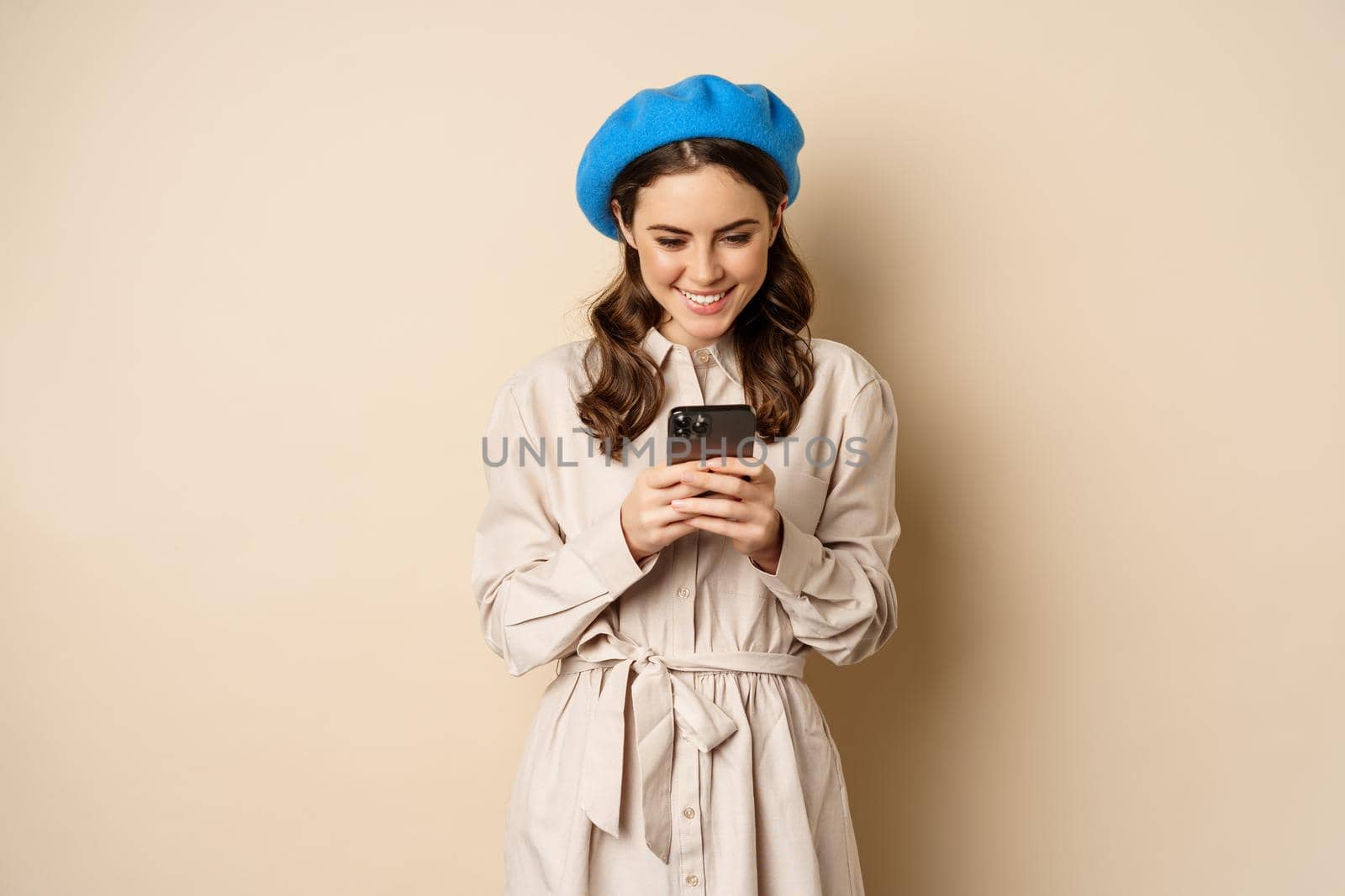 Modern girl using smartphone, smiling and posing happy with mobile phone, standing over beige background by Benzoix